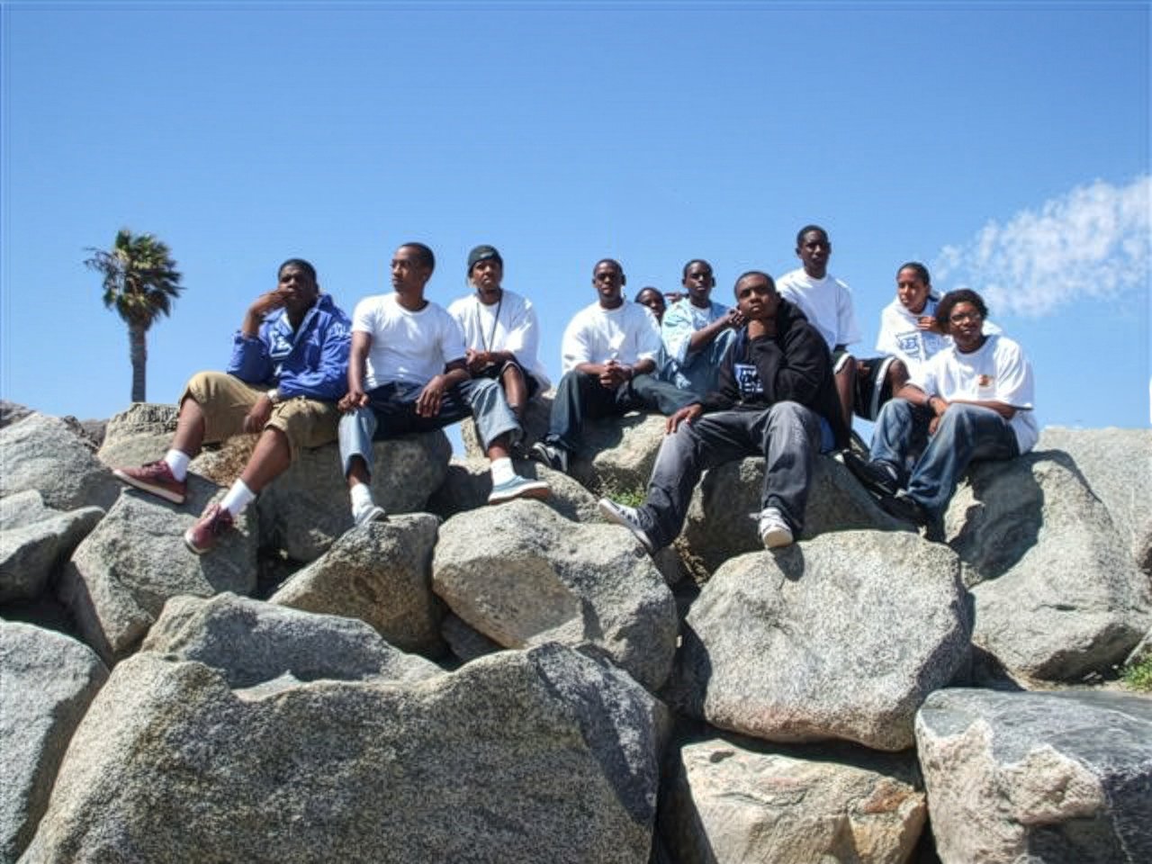 Group of ten young men sitting on large rocks outdoors under a clear blue sky, with a palm tree in the background.