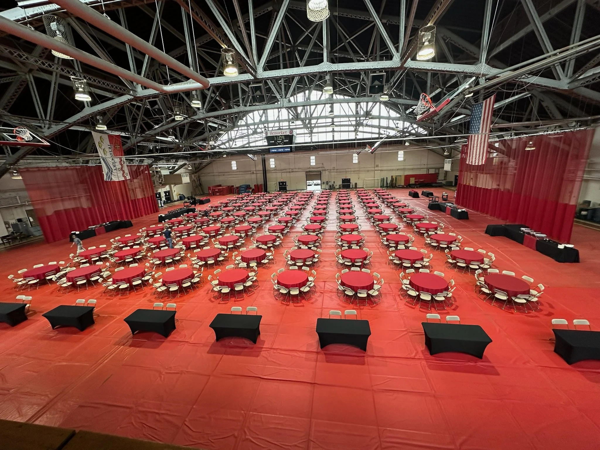 Large indoor event space set up with numerous round tables covered with red tablecloths and surrounded by chairs, with a black table running along the front. The gymnasium has a high metal ceiling with exposed beams and a basketball hoop, and red curtains on the sides.