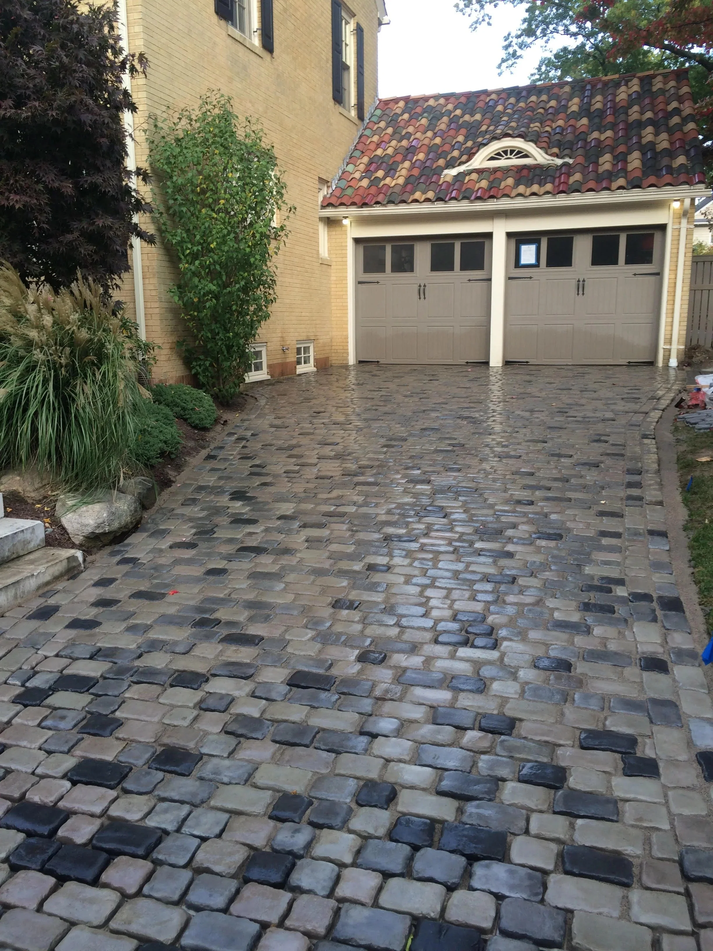 A cobblestone driveway in front of a yellow brick house with a garage door, plants on the left, and trees in the background.
