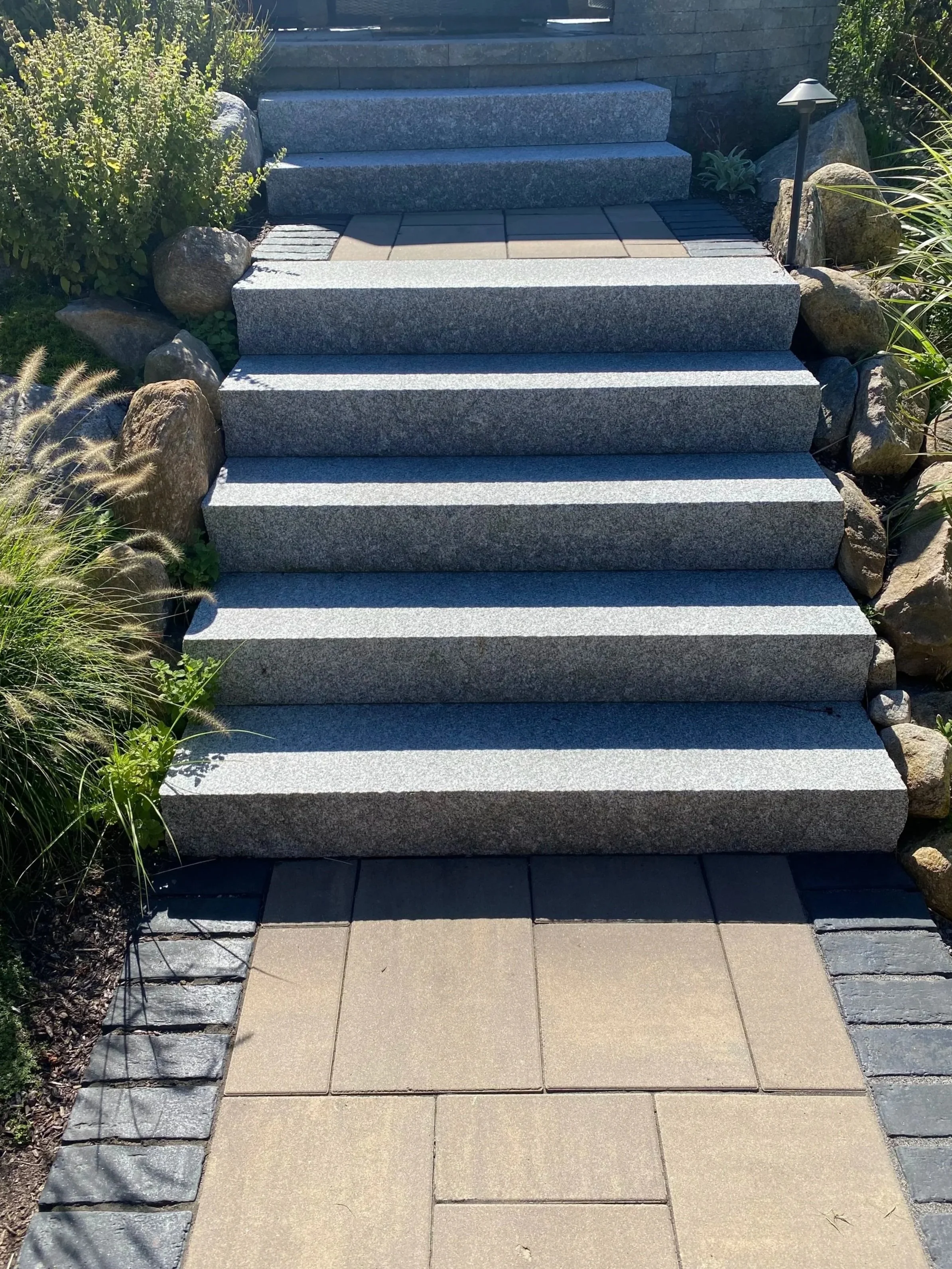 A set of outdoor stairs made of gray granite leading up to a higher level, flanked by landscaped rocks and plants, with lighting fixtures on the side.
