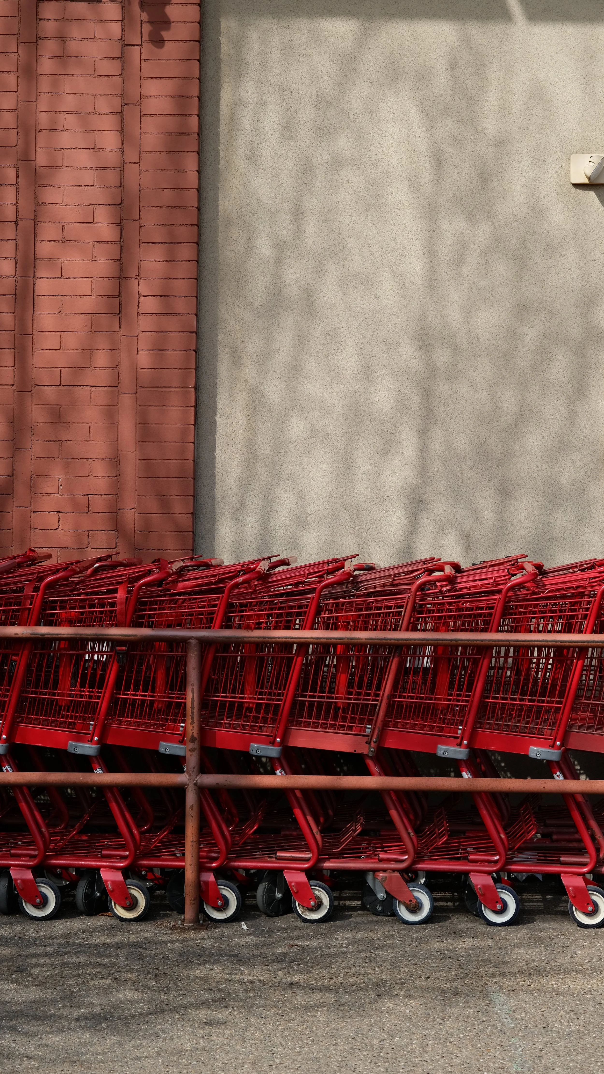 Several red shopping carts lined up and stacked outside against a wall, with a brick wall on the left and a plain gray wall on the right, and shadows of a tree cast on the gray wall.