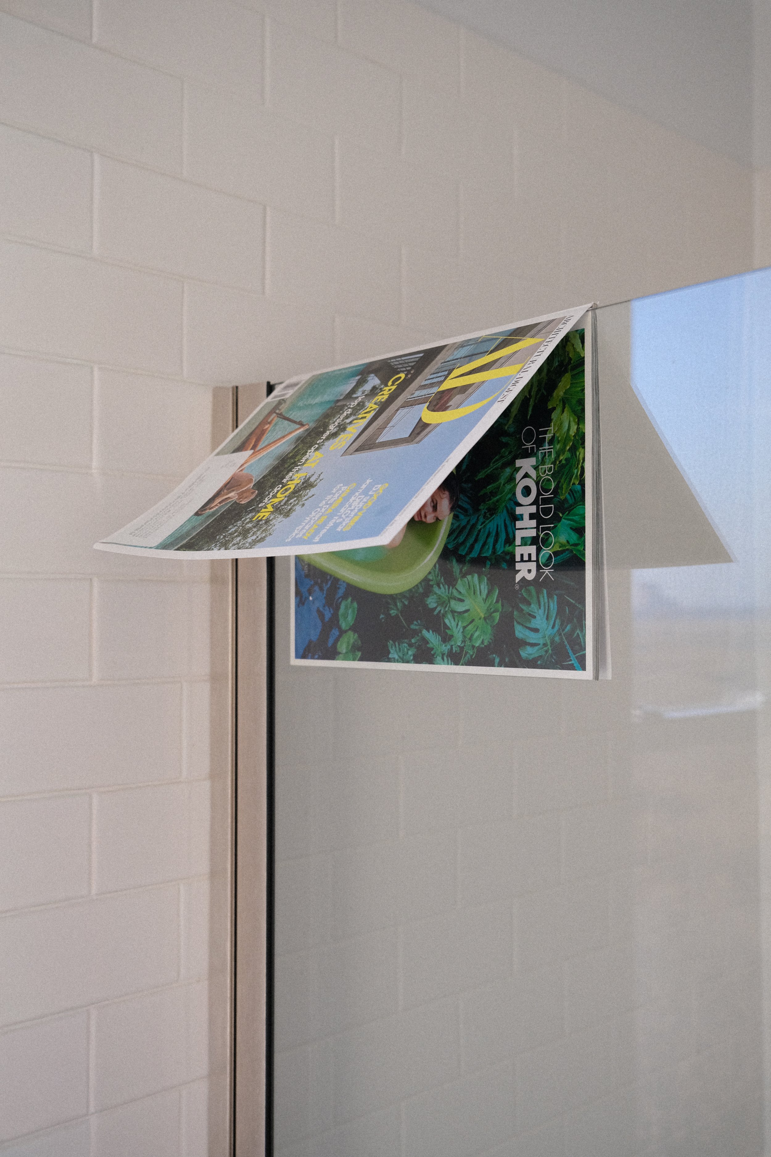 A magazine holder mounted on a wall holds magazines, including one with the title "The Book of Kohler" and another showing a person in a pool.