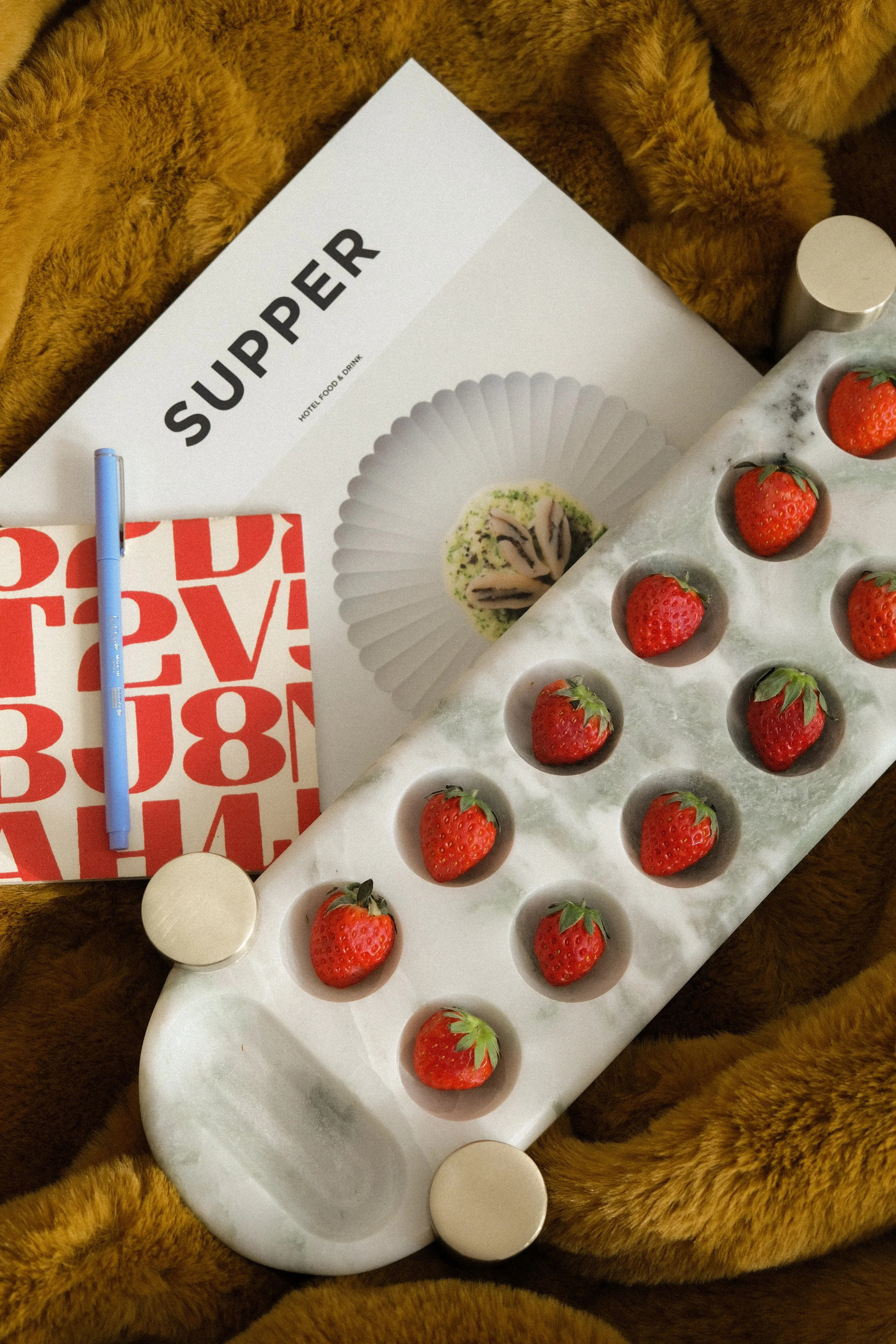 Fresh strawberries on a marble tray, a white and red book, a blue pen, a brochure with food and drink images, and a brown furry blanket.