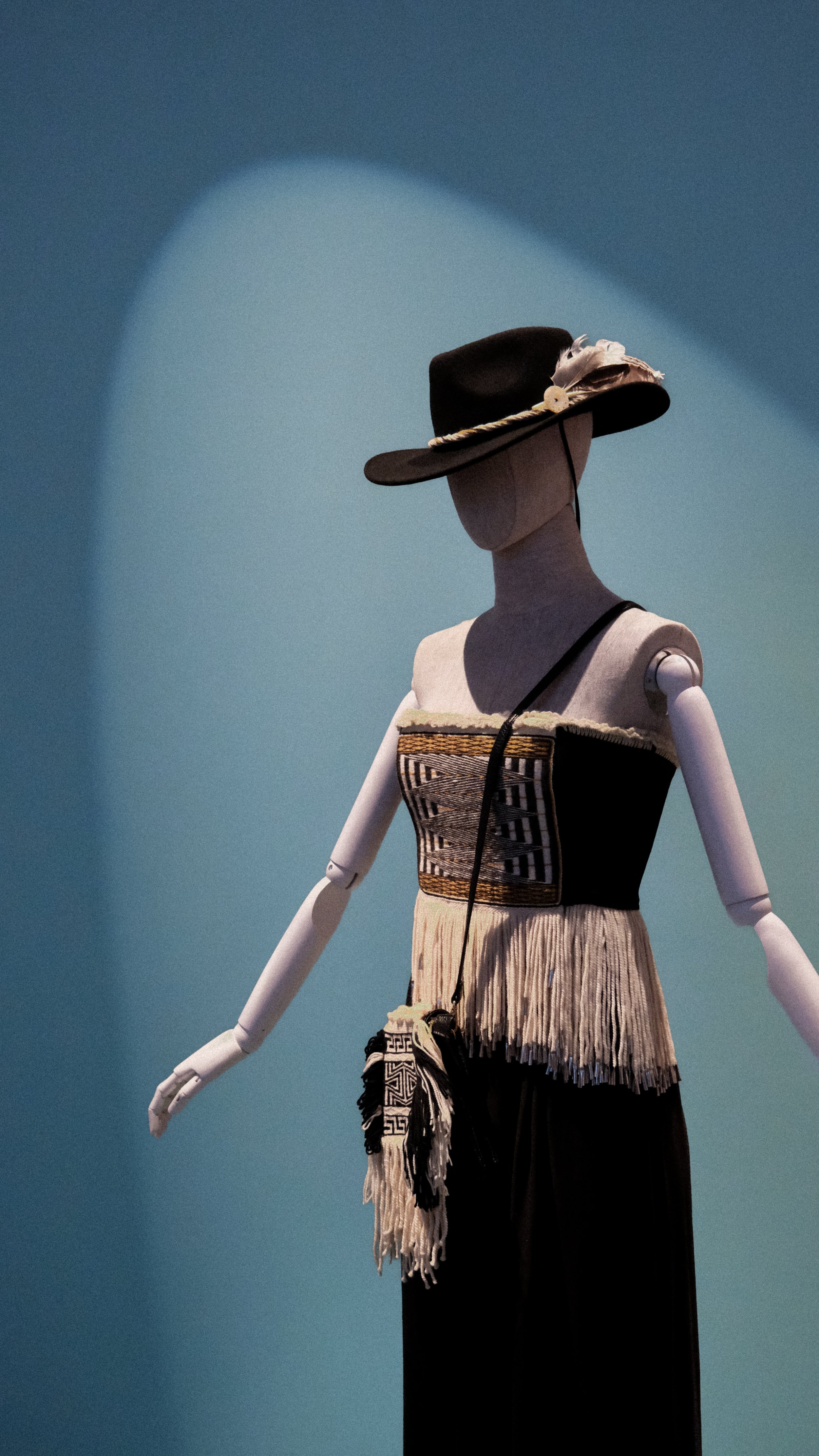 A mannequin dressed in traditional attire with a wide-brimmed hat, a woven top, fringed skirt, and a small shoulder bag, against a blue background.