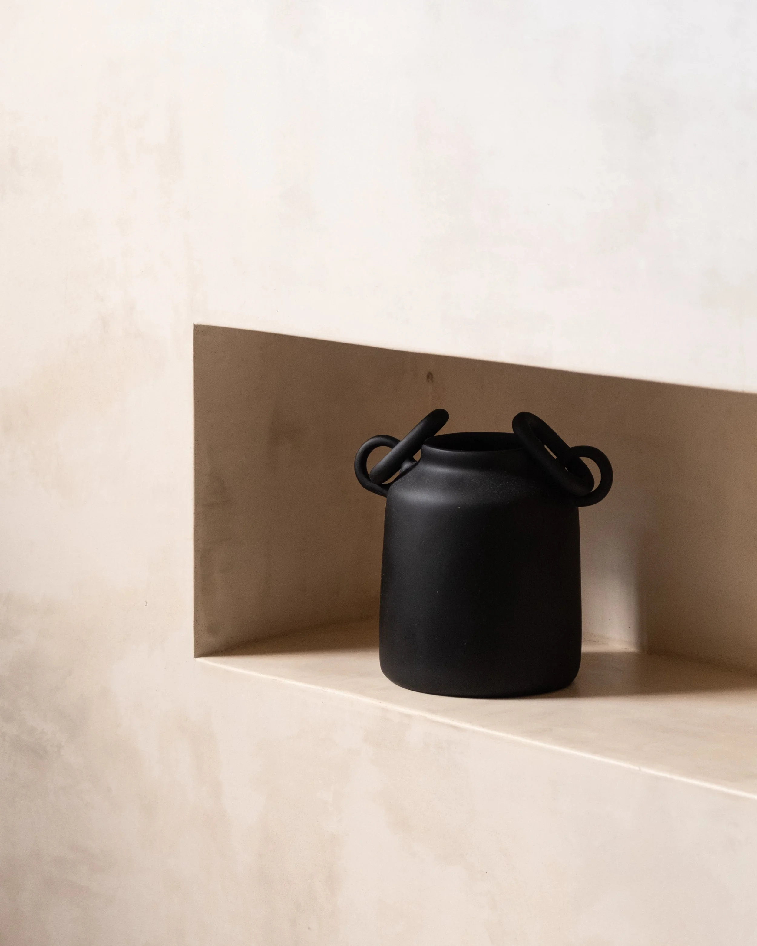 A matte black ceramic vase with looped handles, placed on a beige shelf within a recessed wall niche.