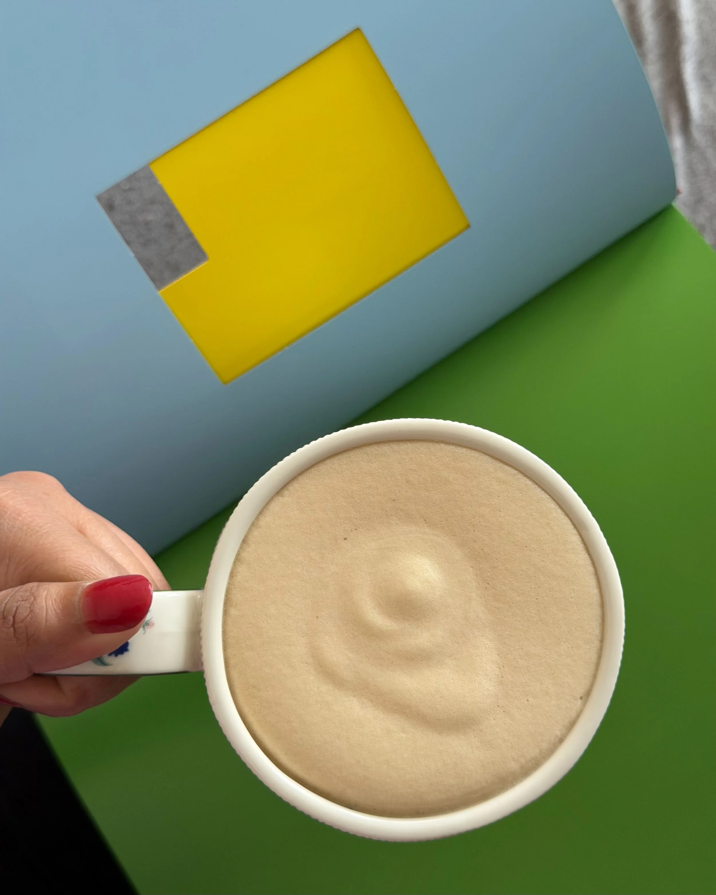 A hand holding a cup of coffee or latte with a ripple pattern on the foam, against a colorful background with blue, green, and yellow surfaces.