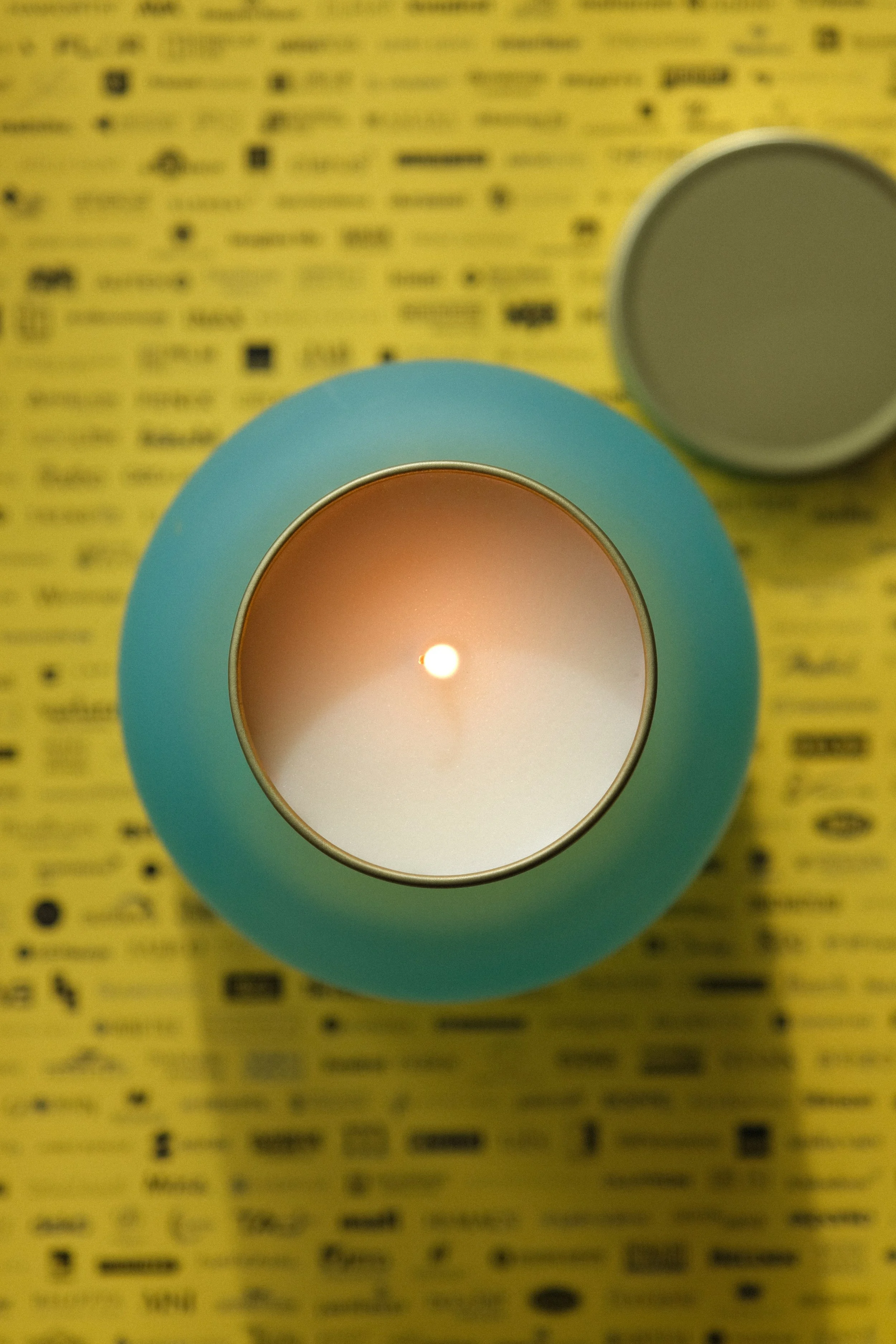 A teal-colored candle holder with a lit white candle inside, placed on a yellow surface with black text and symbols. The candle's lid is removed and placed nearby.