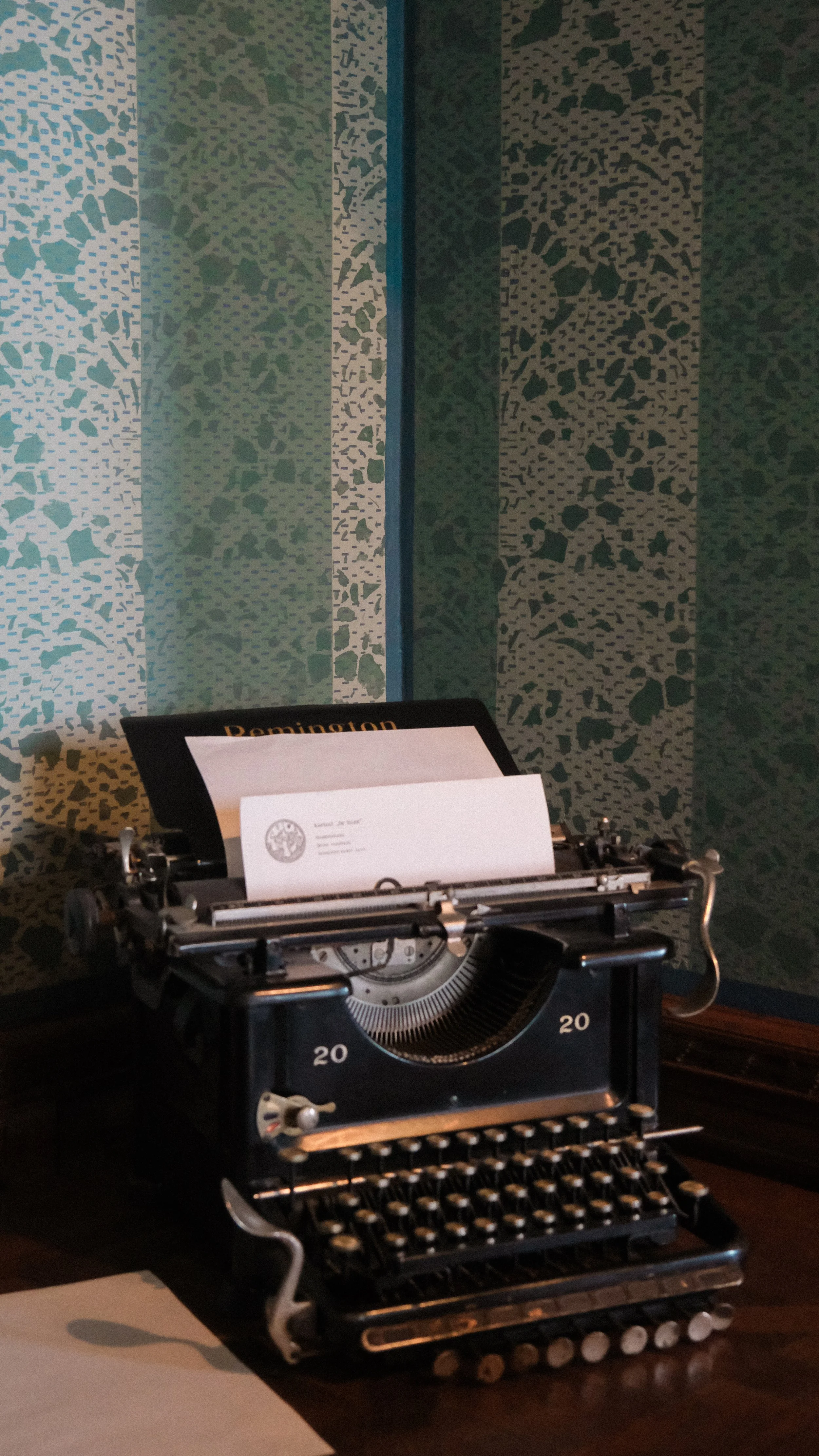 A vintage black typewriter with a sheet of paper inserted, placed on a wooden surface with patterned green and blue wallpaper in the background.