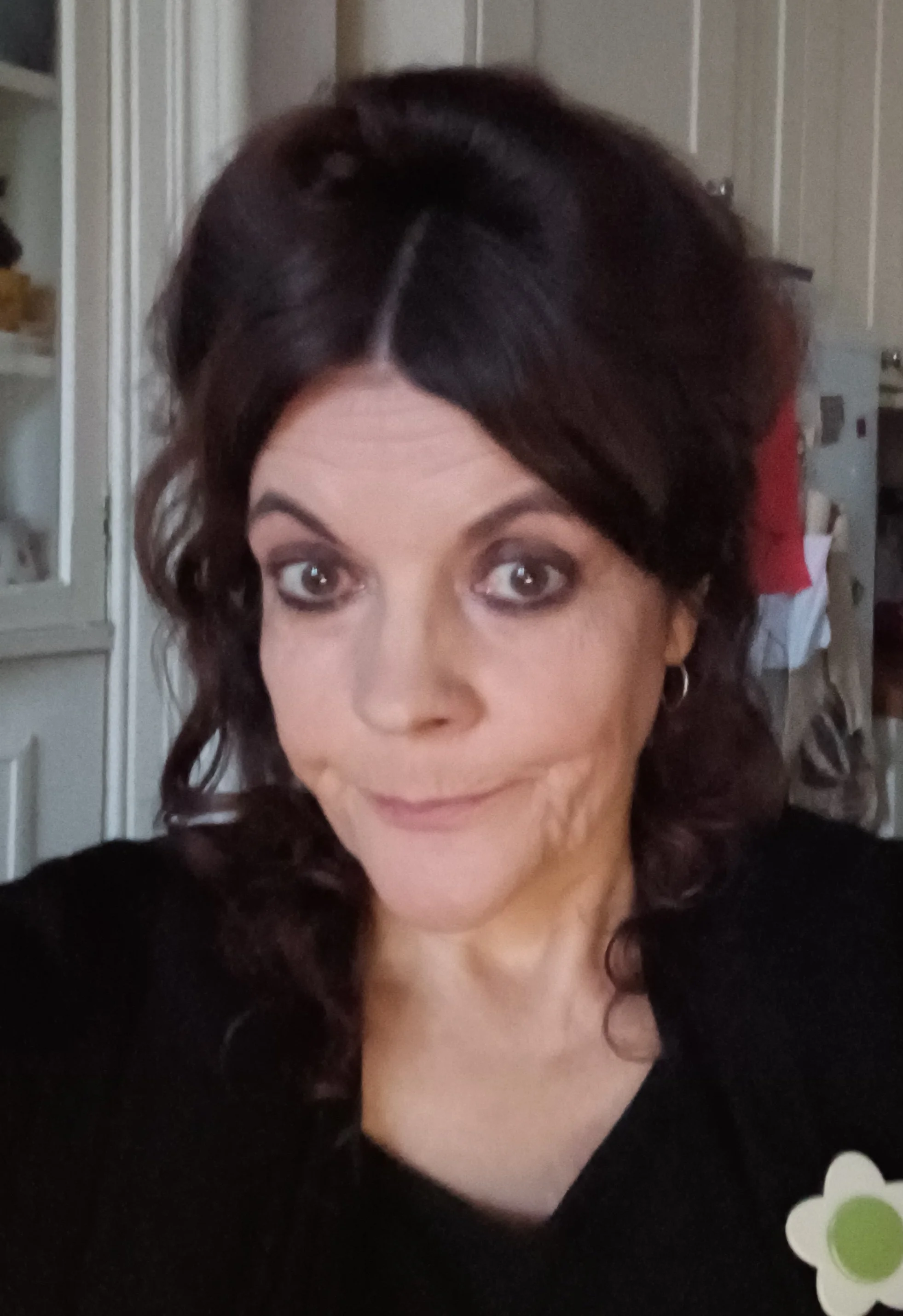 A woman with dark curly hair, wearing makeup, and a black top, facing the camera with a slight smile in a home setting.