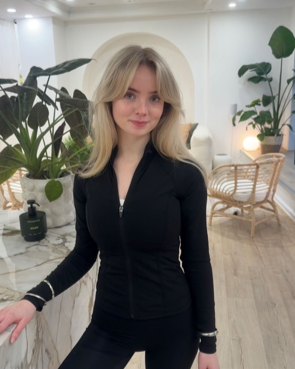 Young woman with blonde hair in a black zip-up top standing in a modern, plant-decorated indoor space.