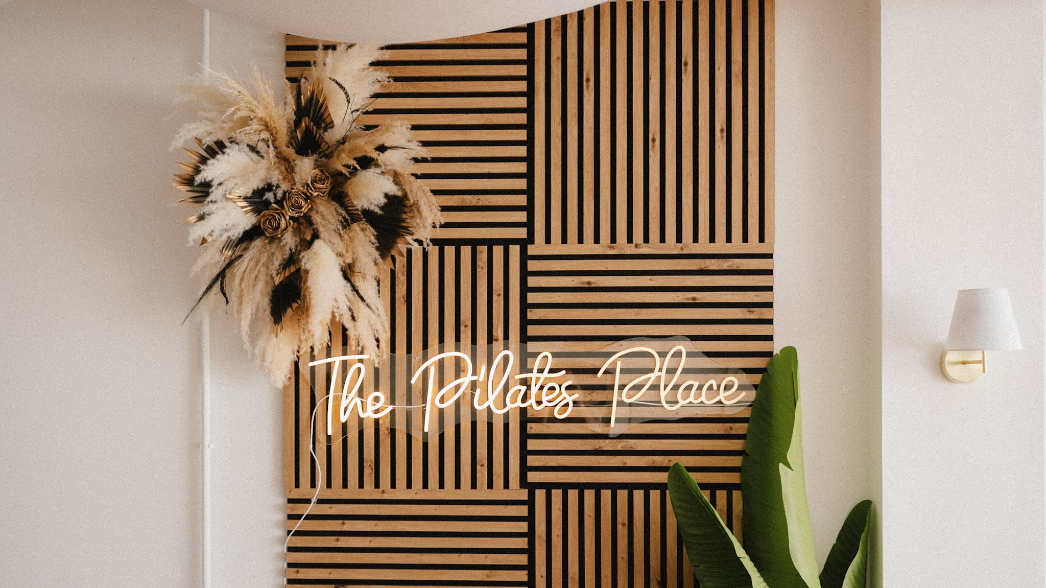 Decorative wall with wooden slats, a floral arrangement with pampas grass, and a neon sign that says 'The Pilates Place,' in a modern interior.