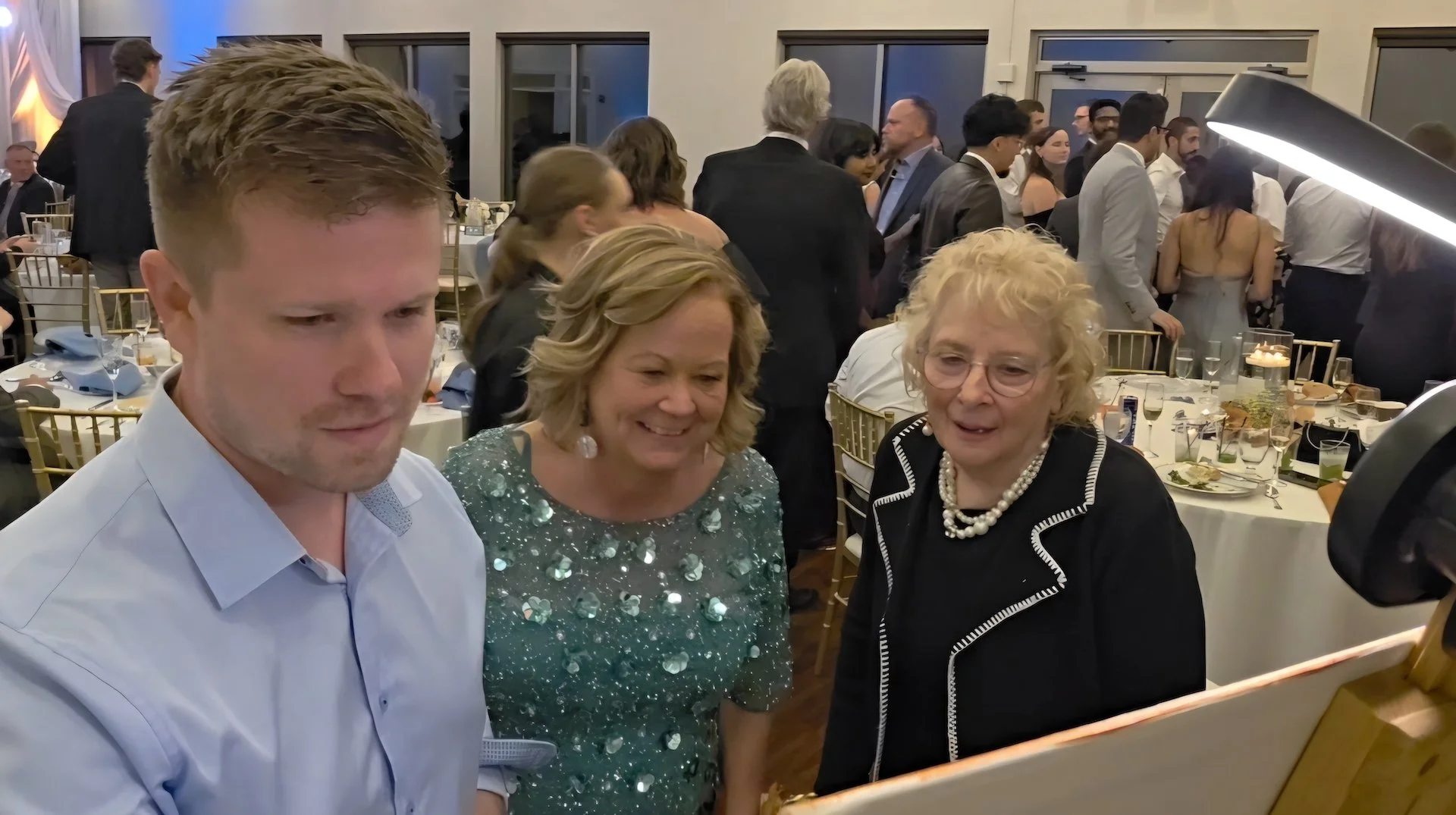 A live wedding painter paints during a wedding reception while two guests watch and smile at his artwork
