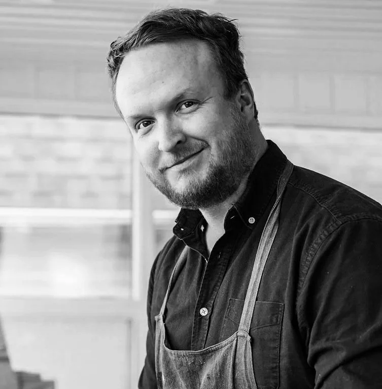 A man with a beard and short hair, wearing a dark shirt and apron, smiling and looking at the camera in a black-and-white photo, with a blurred background.