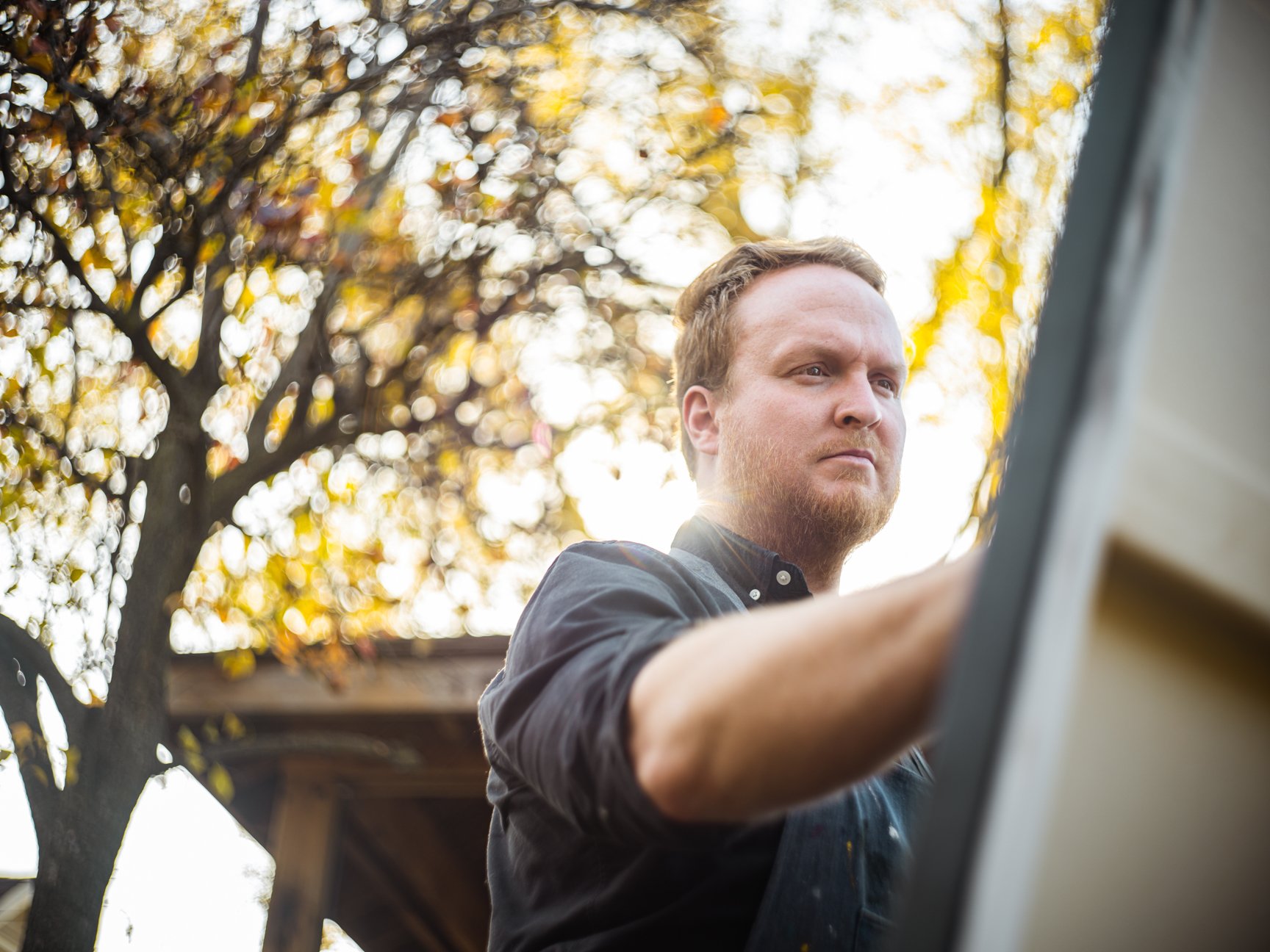 A man with red hair and beard wearing a dark shirt, working on a paintings outdoors during sunset with a tree and a wooden structure in the background.