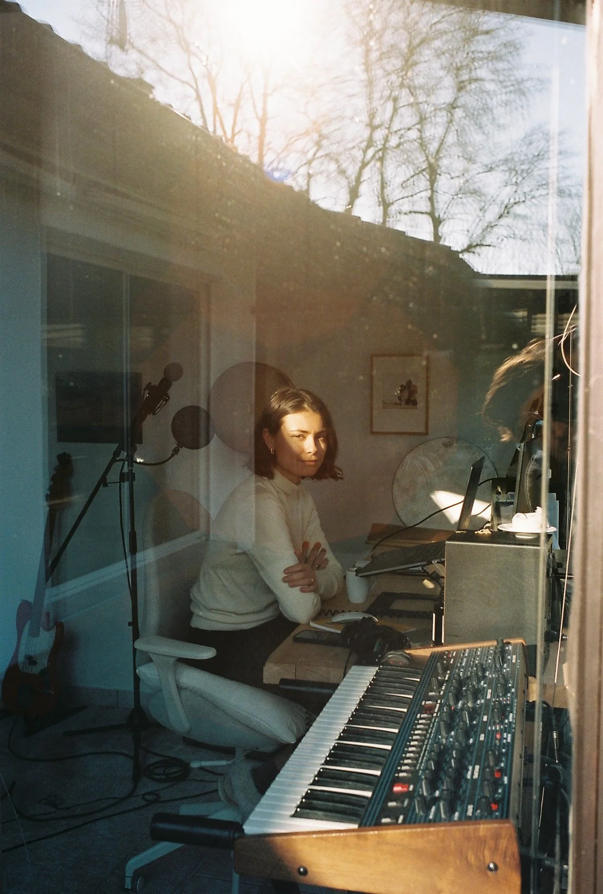 A woman sitting at a music studio desk, with a synthesizer keyboard in the foreground, a computer monitor, and audio equipment. The scene is captured through a glass window with reflections of daylight and trees outside.