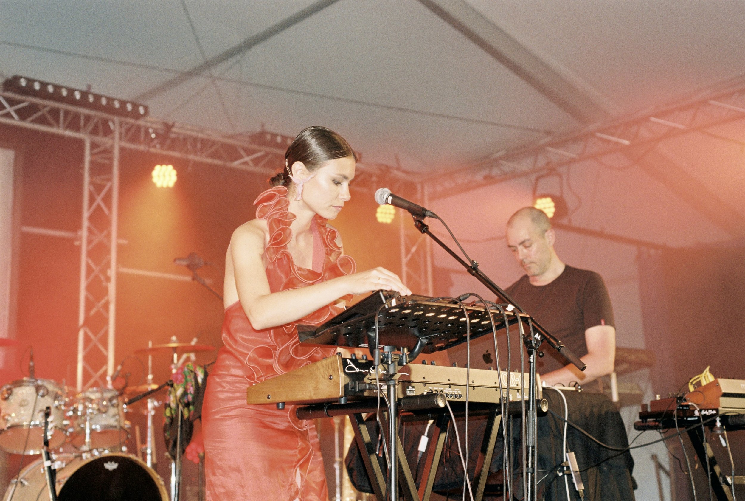 A female musician in a red dress playing a keyboard with a male musician in black playing another keyboard in the background on a stage with orange lighting.