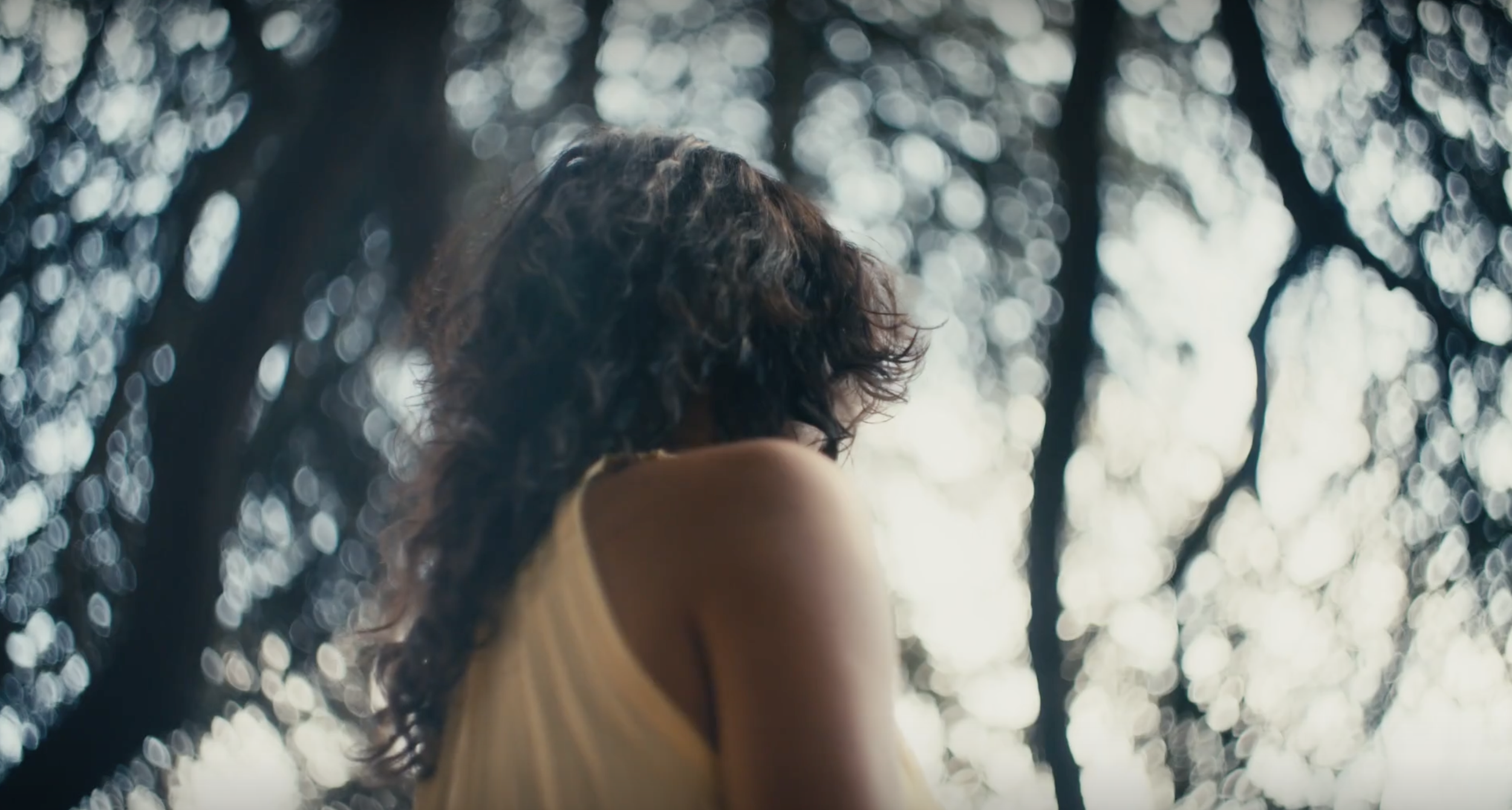 A woman with curly hair wearing a yellow top, looking up in a forest with sunlight filtering through the trees, creating a bokeh background.