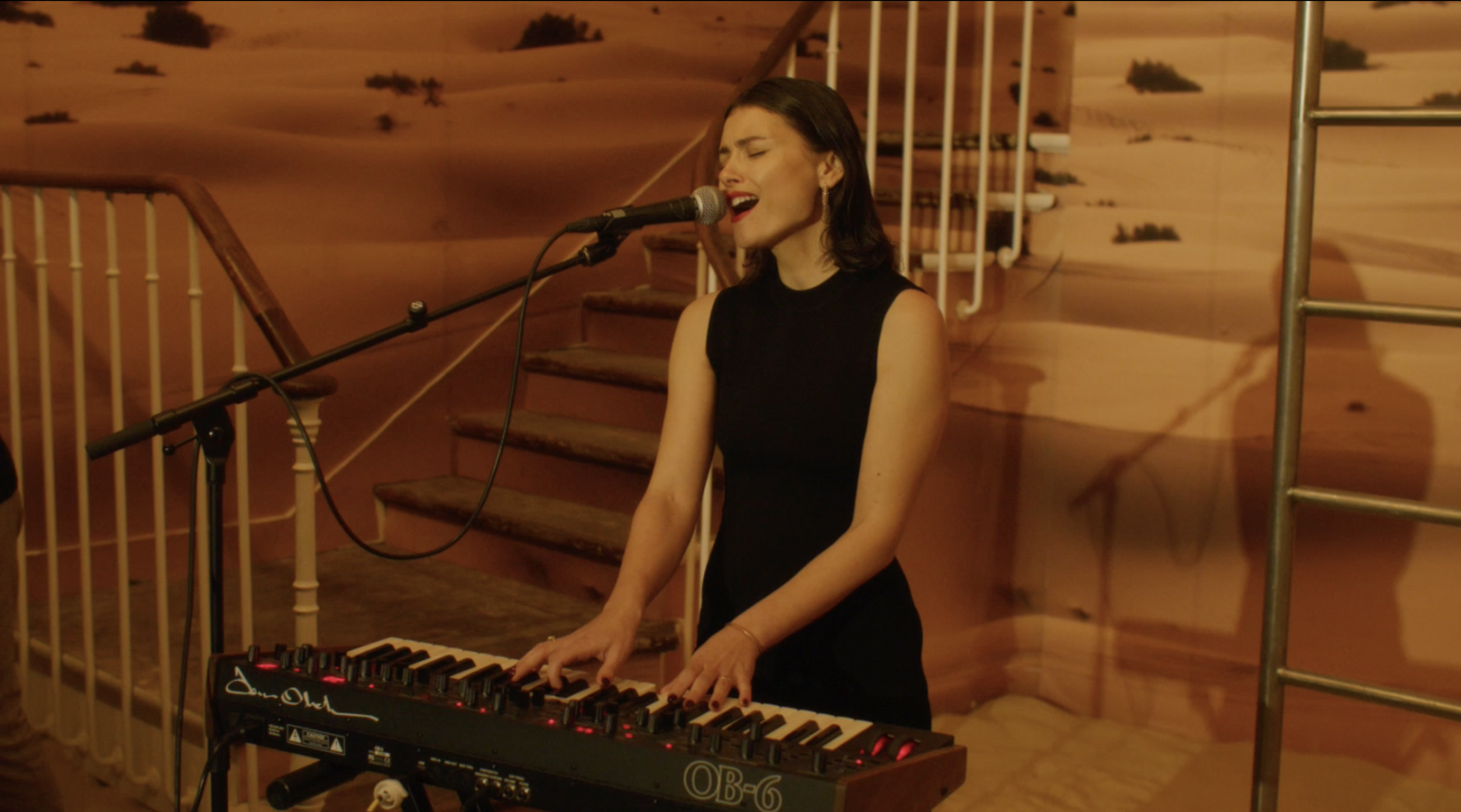 A woman in a black sleeveless dress is singing and playing a keyboard, standing in front of a microphone. The background depicts a desert scene with sand dunes and plants.