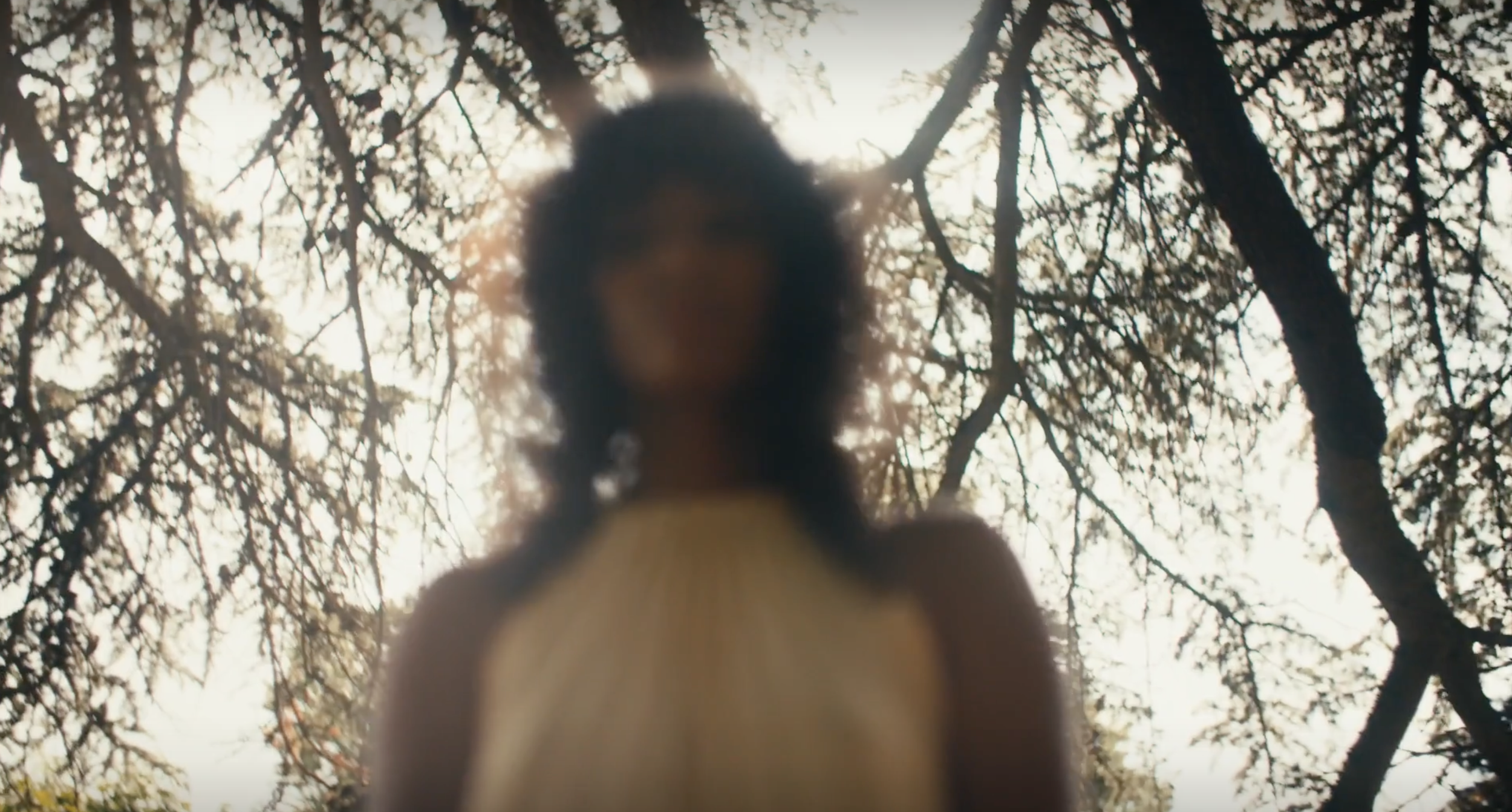 A woman standing outdoors in a forest with sunlight filtering through the trees, viewed from below.