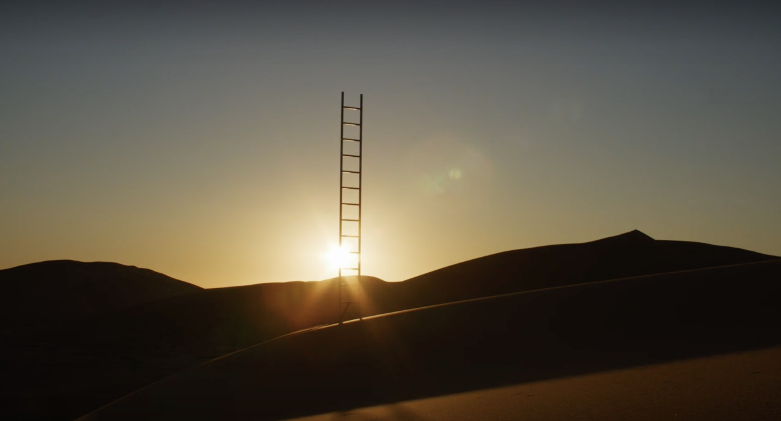 Silhouette of a ladder standing on sand dunes during sunset, with the sun low on the horizon.