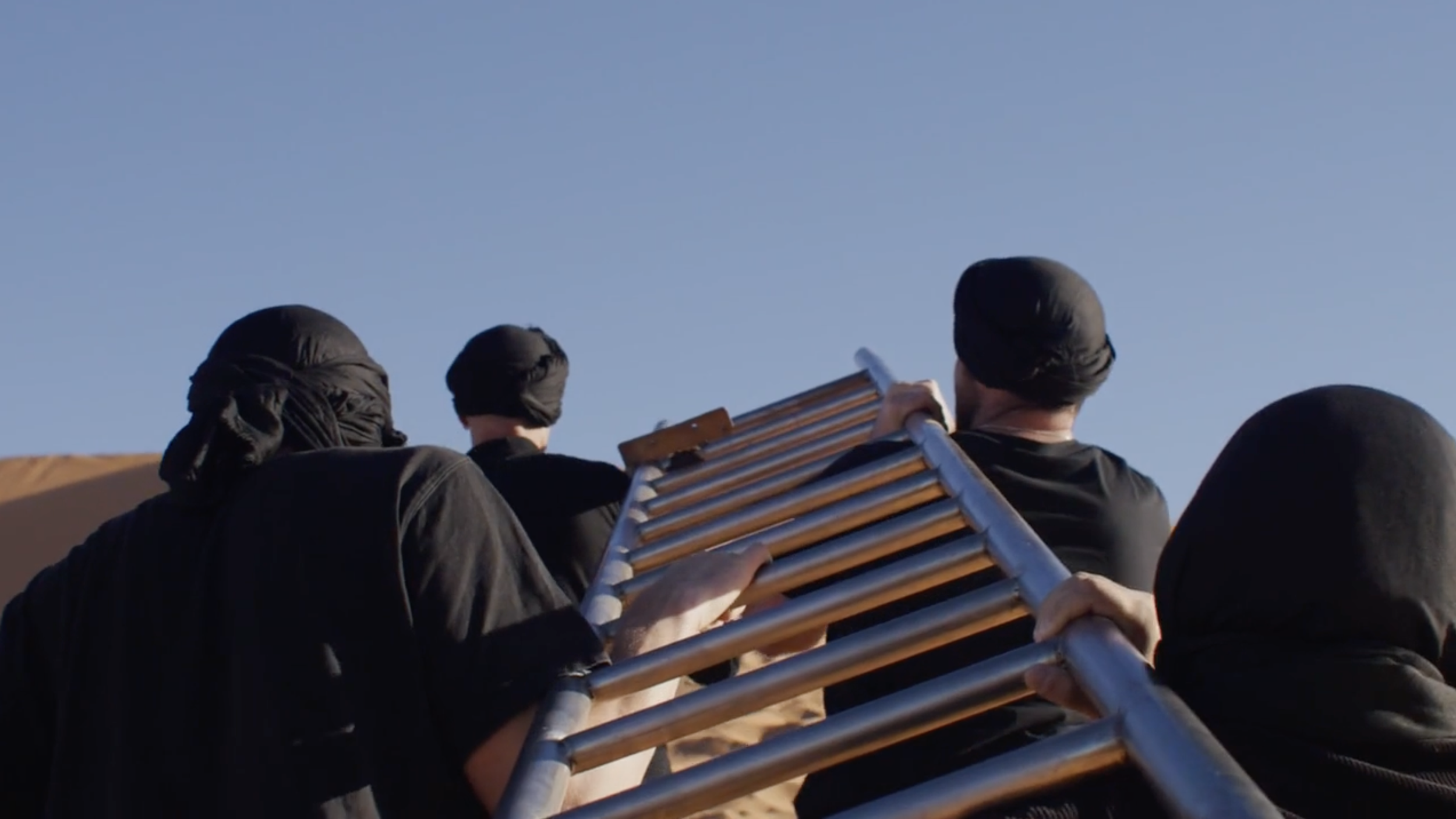 People climbing a metal ladder on sand dunes, all wearing black clothing and head coverings, against a clear blue sky.