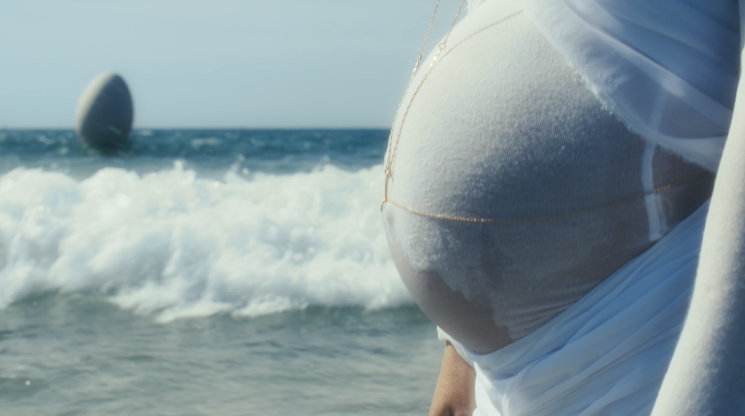 Close-up of a woman on the beach wearing a white bikini top and a necklace, with the ocean and a sailboat in the background.