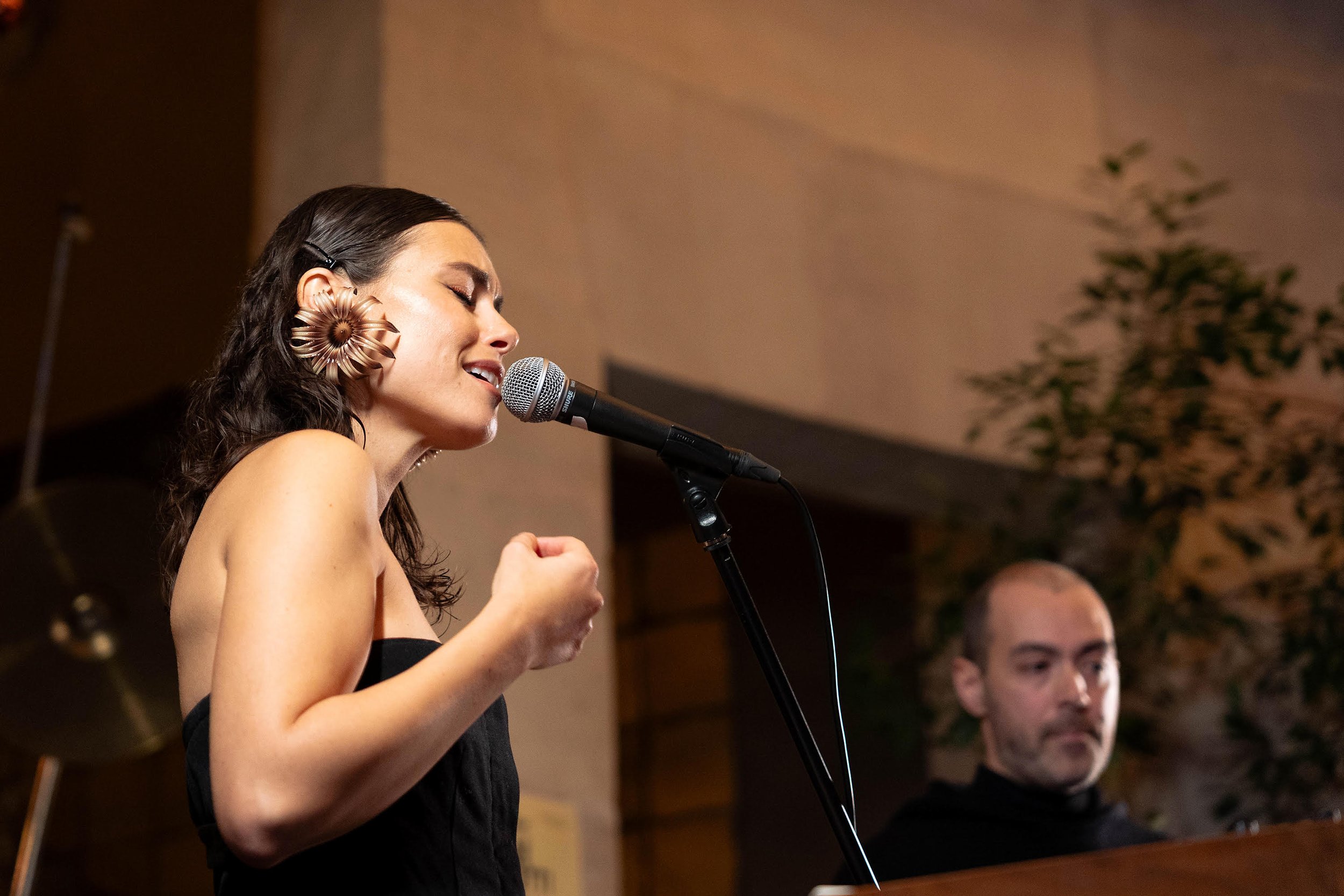 A woman passionately singing into a microphone with her eyes closed and a clenched fist, wearing a black strapless dress and large floral earrings. A man is in the background playing a keyboard, with a neutral expression, wearing a black shirt, and behind them is a beige wall with green foliage.