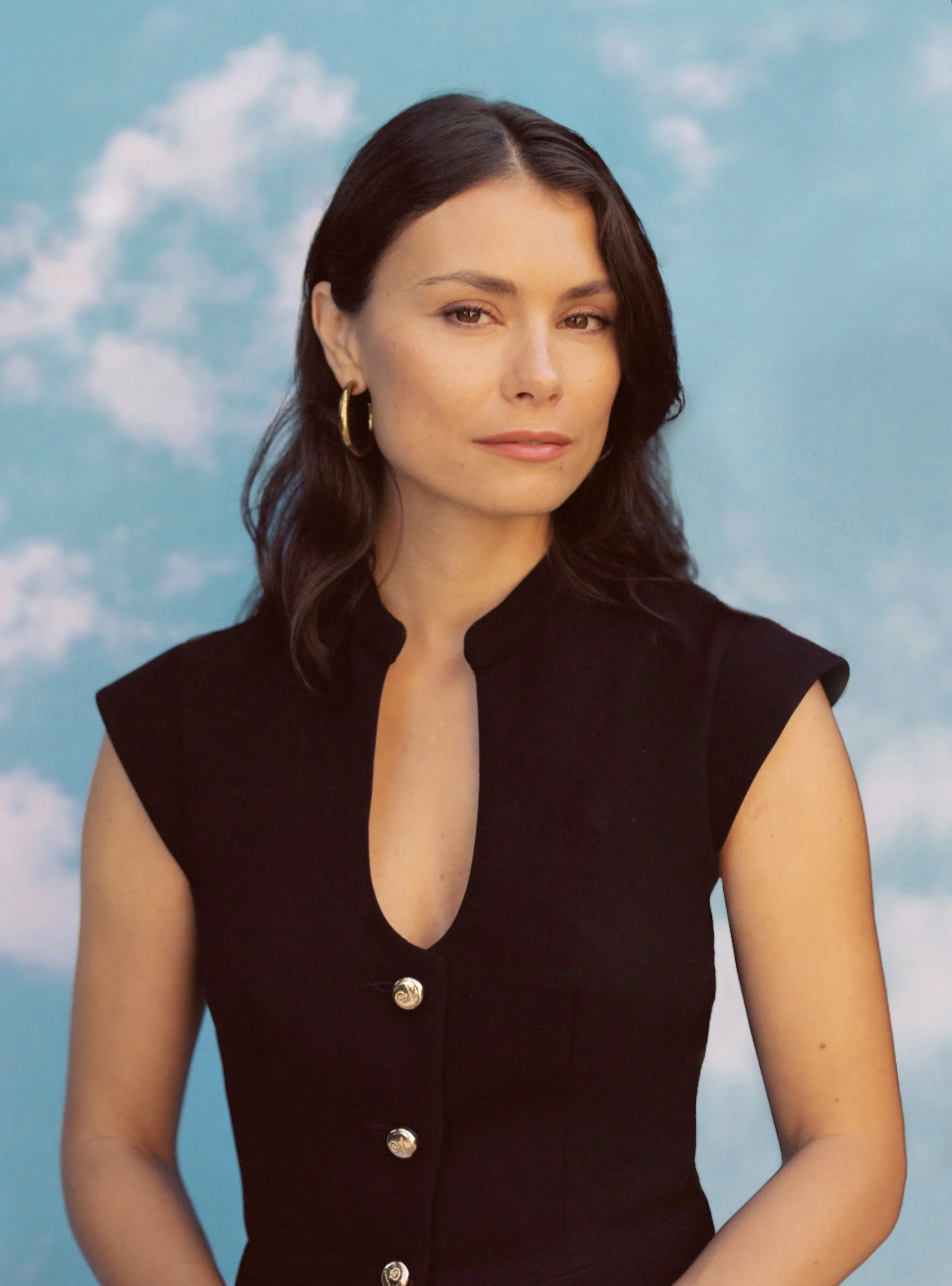 A woman with dark brown hair wearing a black sleeveless top with gold buttons, standing against a blue sky with white clouds background.