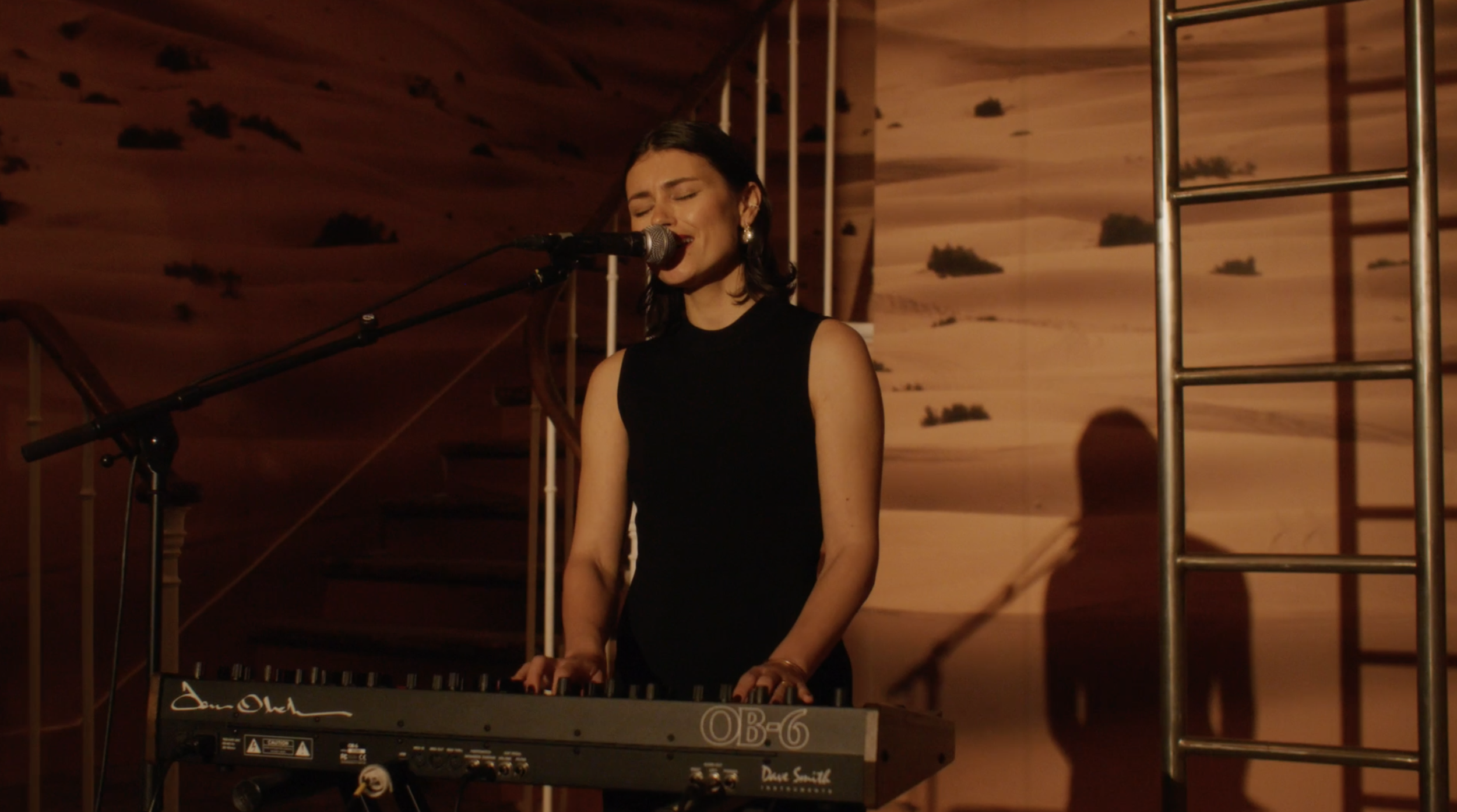 A woman in a black sleeveless dress performing on stage with a keyboard, singing into a microphone, with warm lighting and a backdrop of a desert landscape.