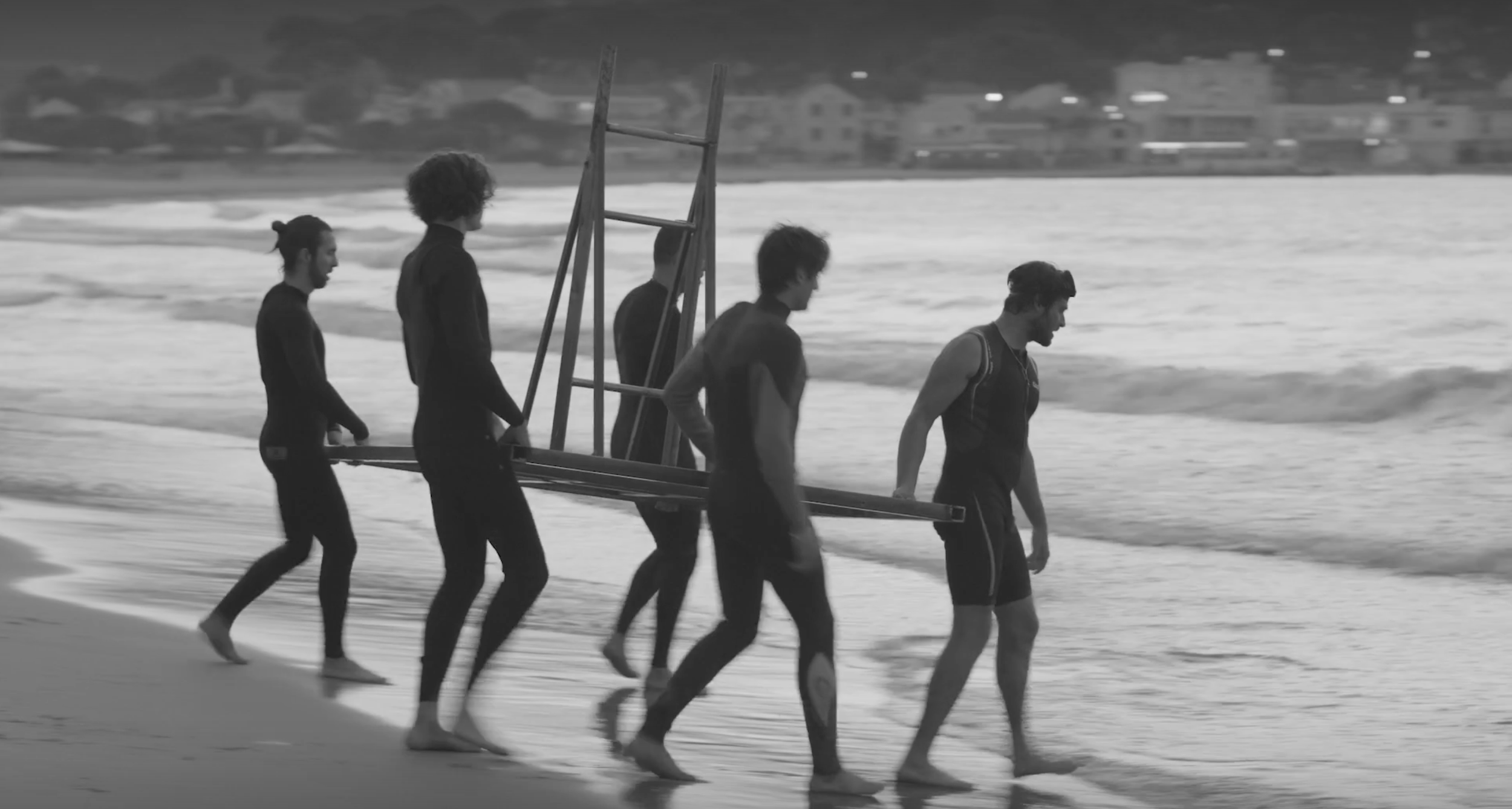Group of five young men walking along the beach carrying a ladder or similar structure towards the water, with waves and houses in the background.