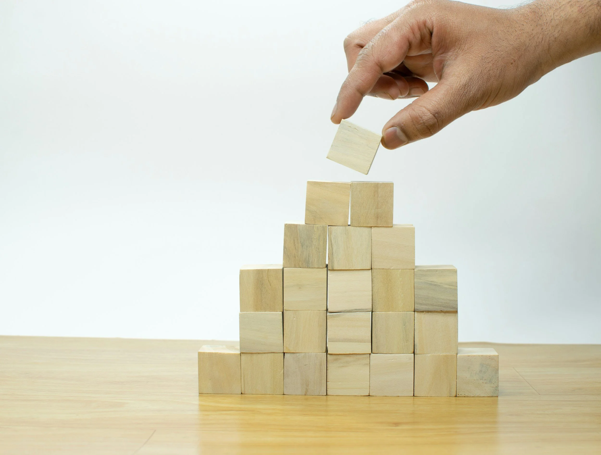 A hand placing a wooden block on top of a stack of wooden blocks arranged in a pyramid shape on a wooden surface.
