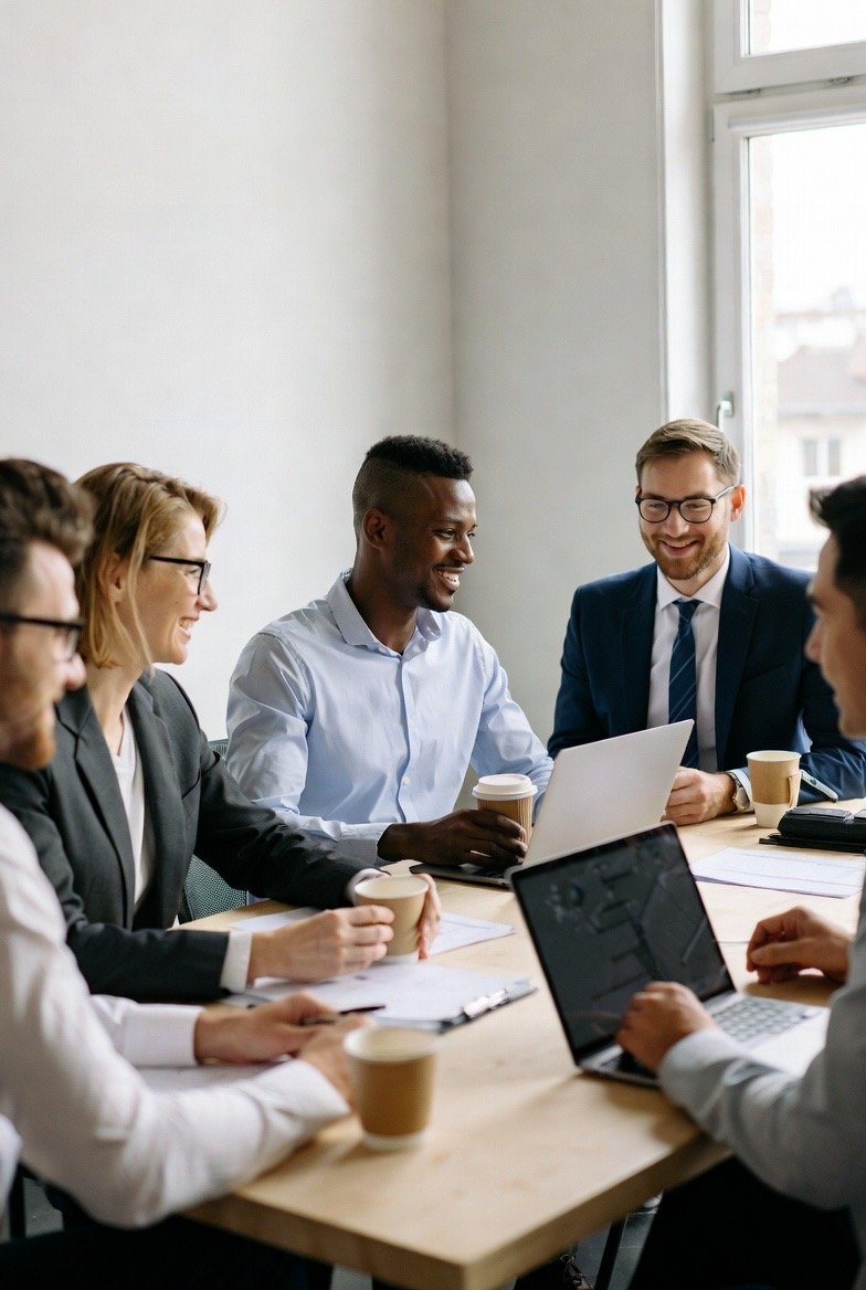 A diverse group of six professionals, three men and three women, sitting around a conference table in a modern office, actively engaged in a meeting, smiling, and holding coffee cups and laptops.