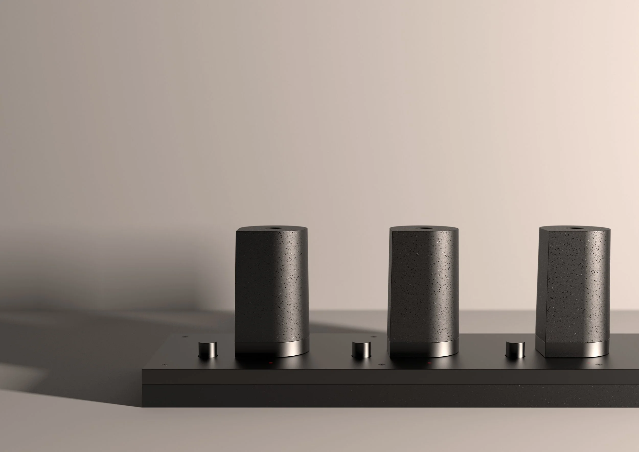 Drei schwarze Lautsprecher auf einem schwarzen Regal, mit je einem Regler und einer kleinen roten Lichtanzeige darunter, vor einem hellen, neutralen Hintergrund.