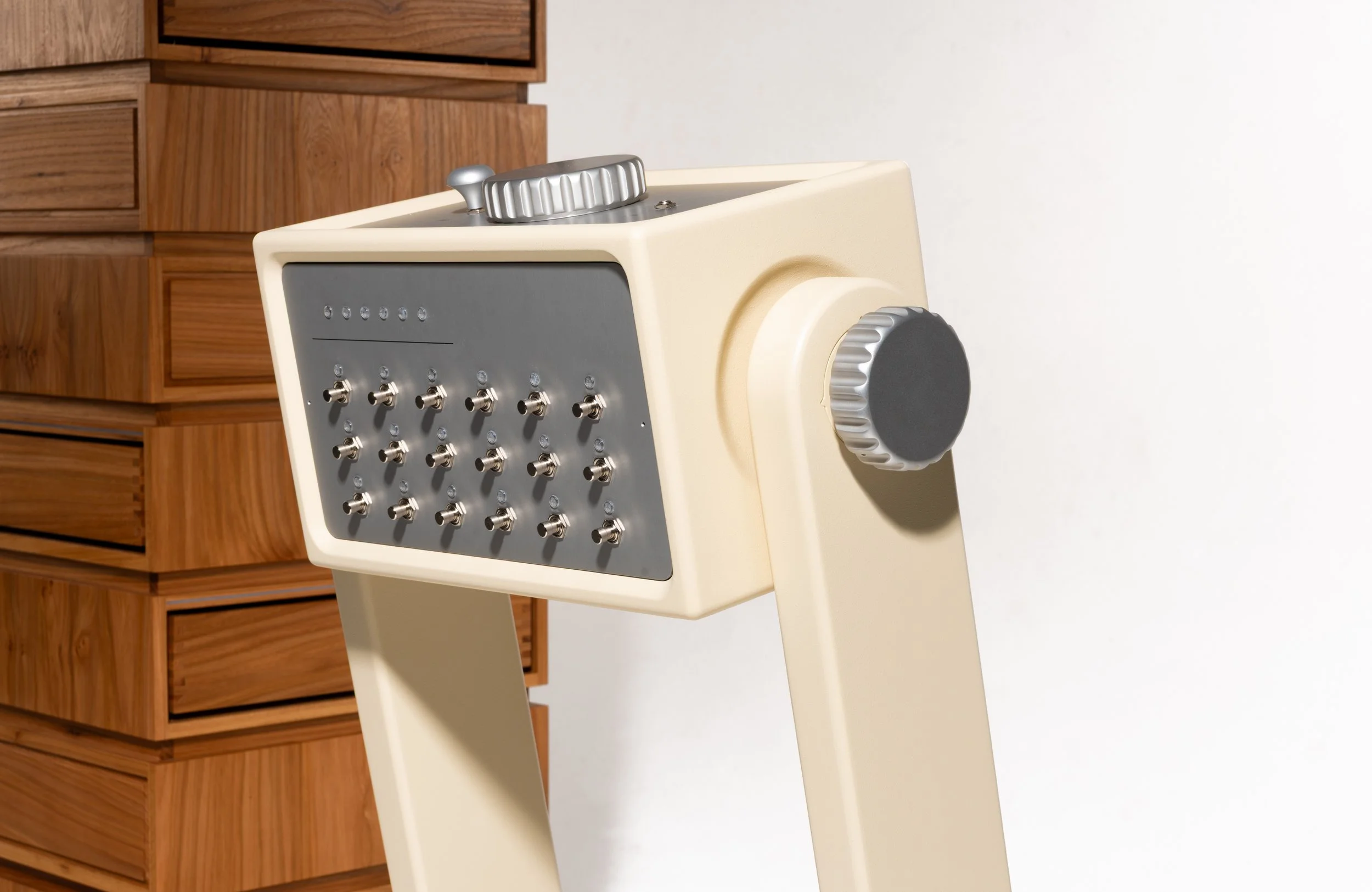 Ein Onkel mit einem grauen Gehäuse und vielen kleinen metallischen Kugeln oder Köpfen auf der Vorderseite, steht neben einem weißen Sockel mit einem großen Riemenrad, im Hintergrund eine Holzkommode.