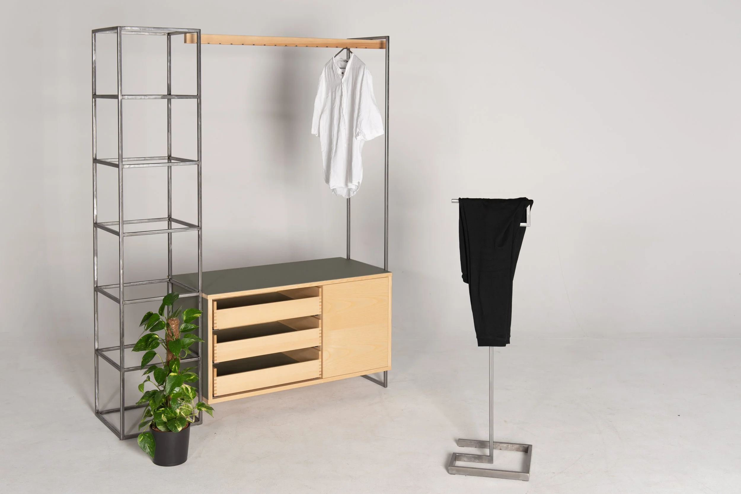 Moderner Kleiderständer mit weißen Hemd, schwarzer Hose und Pflanze in einem minimalistischen Raum.