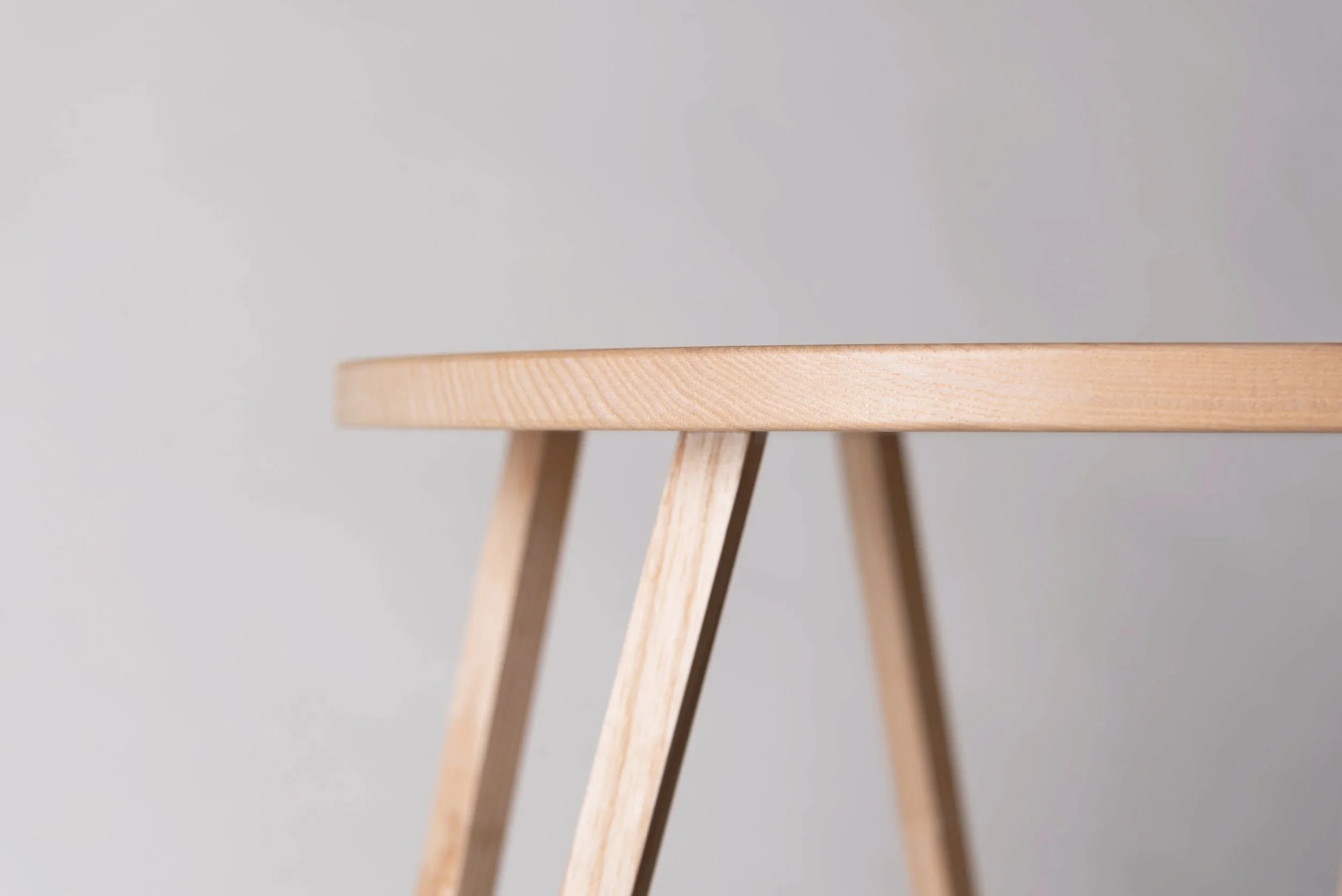 Nahaufnahme eines runden, hölzernen Tisches mit drei sichtbaren Beinen auf einem hellgrauen Hintergrund.