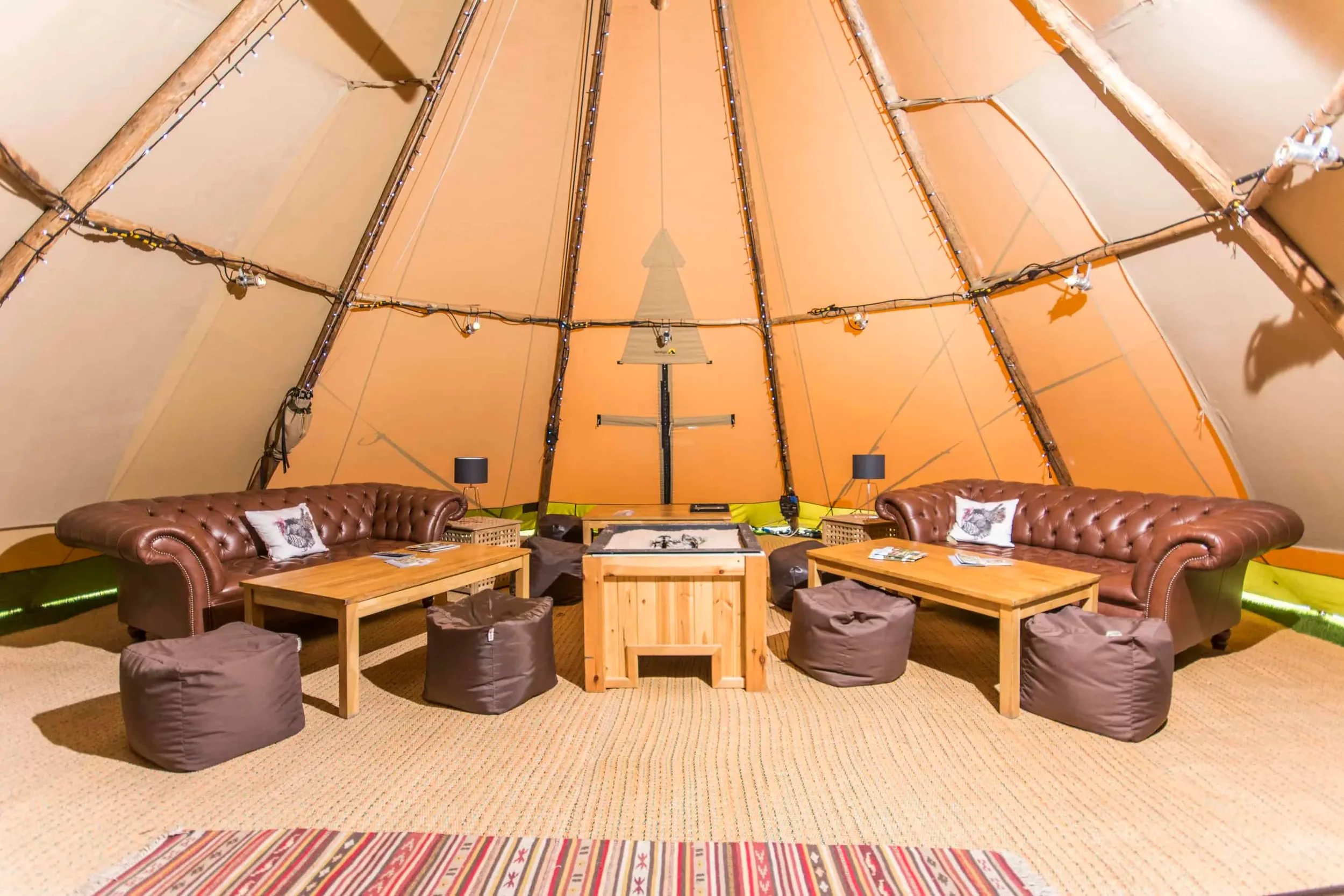 Interior of a tipi tent with two brown leather tufted sofas, wooden tables, brown bean bags, small black lamps, and string lights on the ceiling.