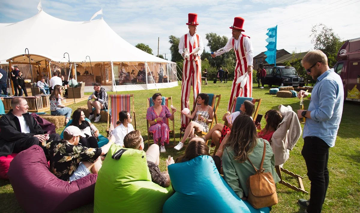 People attending an outdoor event with a circus performance, two entertainers on stilts in striped costumes and tall red hats, and a group seated on colorful bean bags and chairs, enjoying the show.
