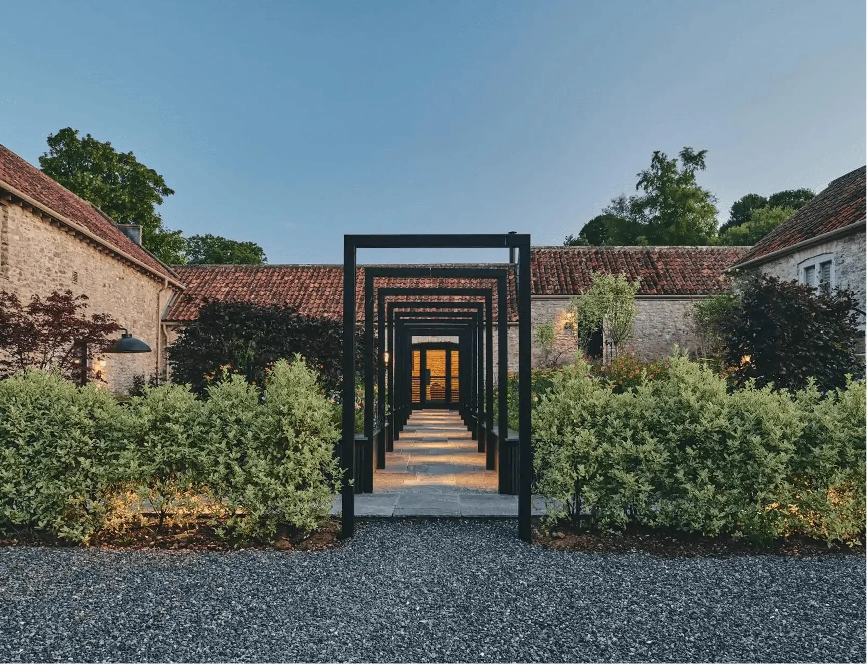 A pathway through a courtyard lined with landscaped bushes, leading to a doorway. The courtyard is surrounded by stone buildings with red tile roofs under a clear blue sky.