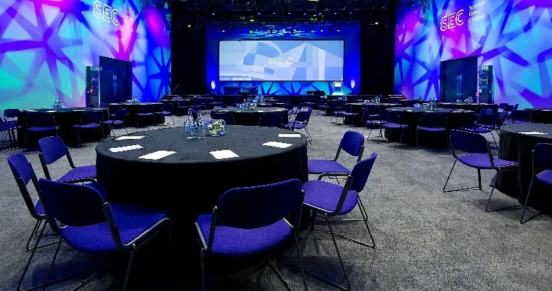 Conference room with round tables, chairs, water bottles, glasses, and notes, set up for a presentation or event, with a large screen and colorful lighting effects.