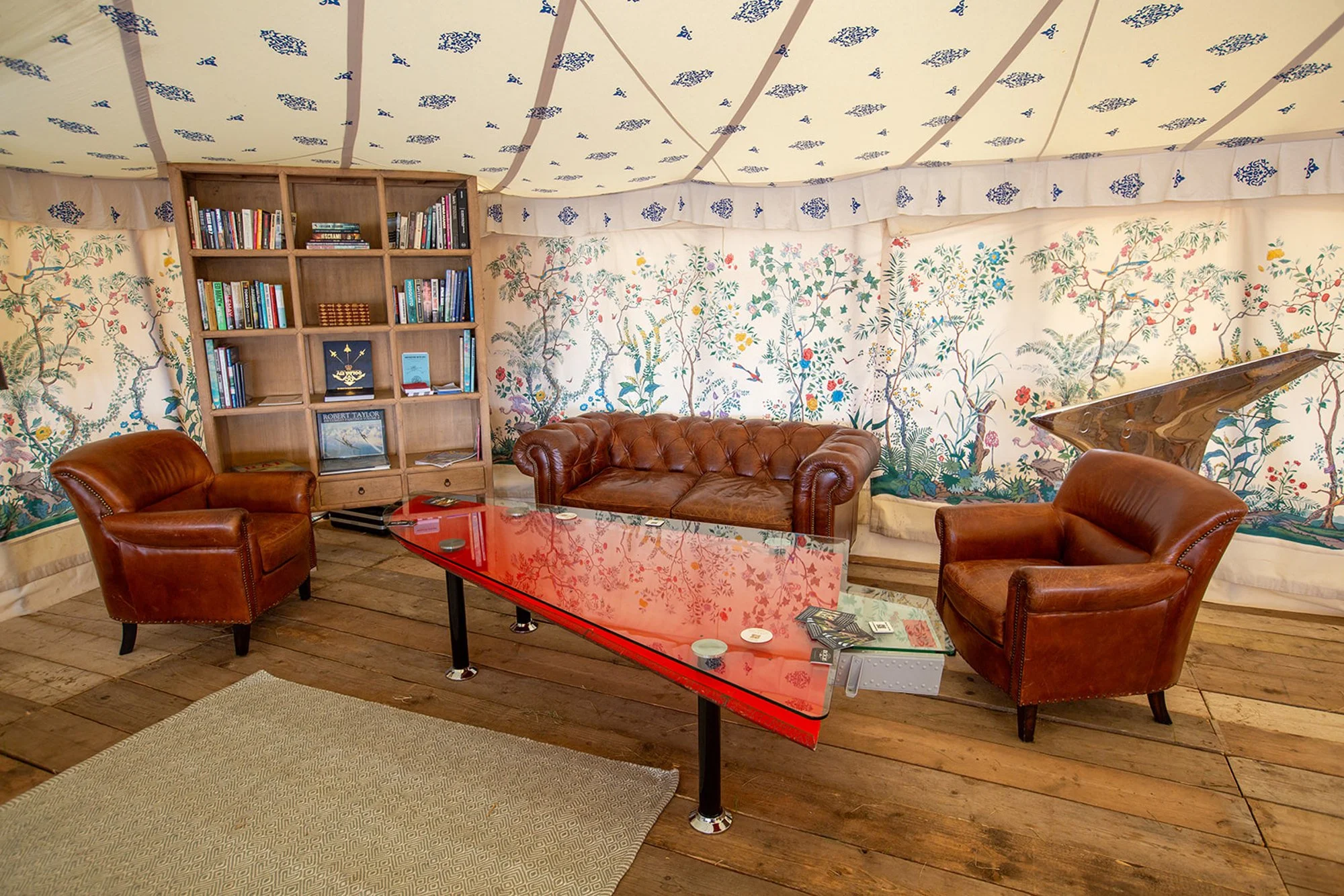 A cozy sitting area inside a tent with floral wallpaper, a wooden bookshelf filled with books, two leather armchairs, a leather sofa, a red glass coffee table, a grand piano, and a beige rug on wooden floor.