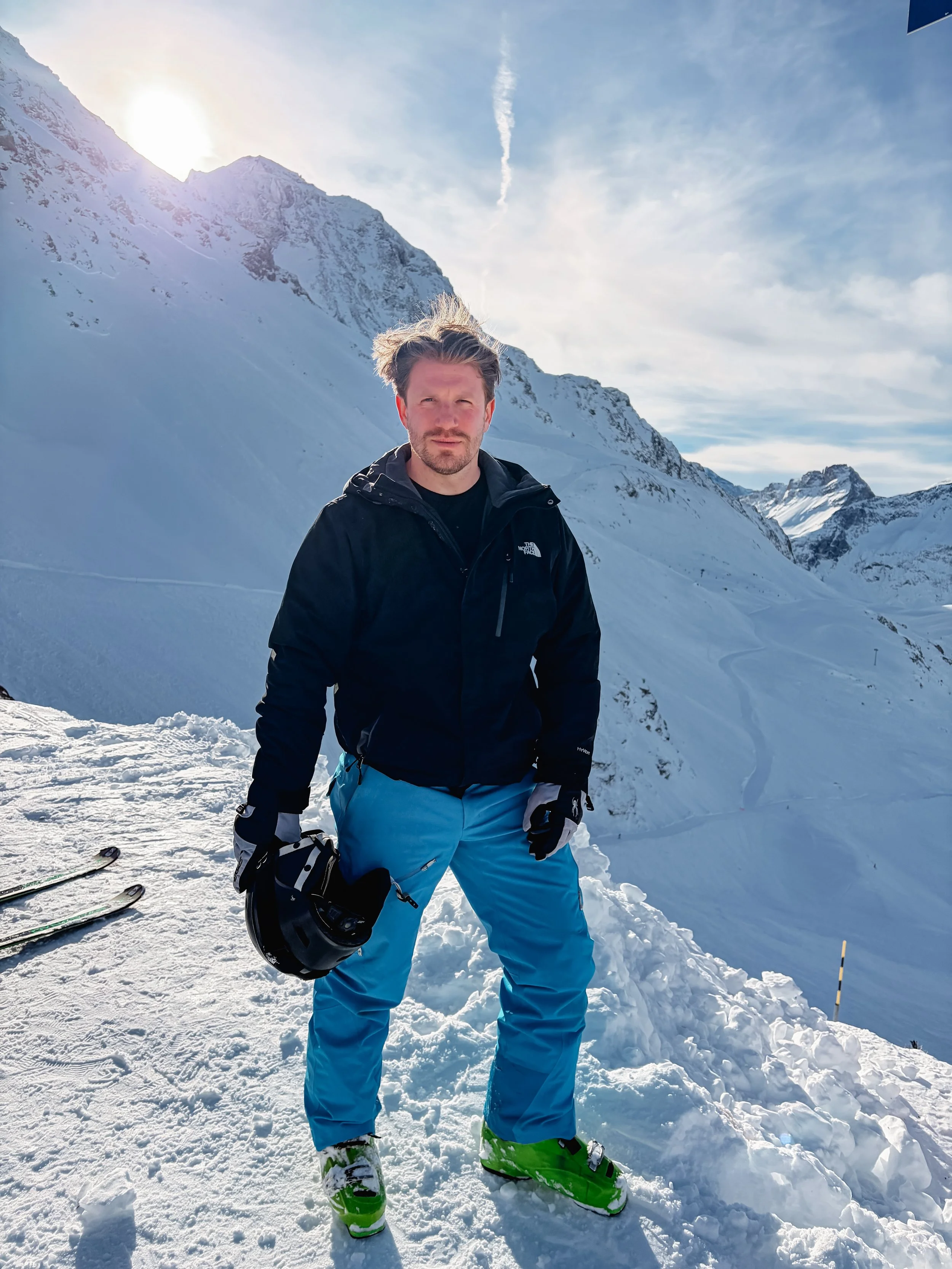 Man standing on snow-covered mountain slope with ski gear, holding a helmet, with snow-capped mountains and a cloudy sky in the background.