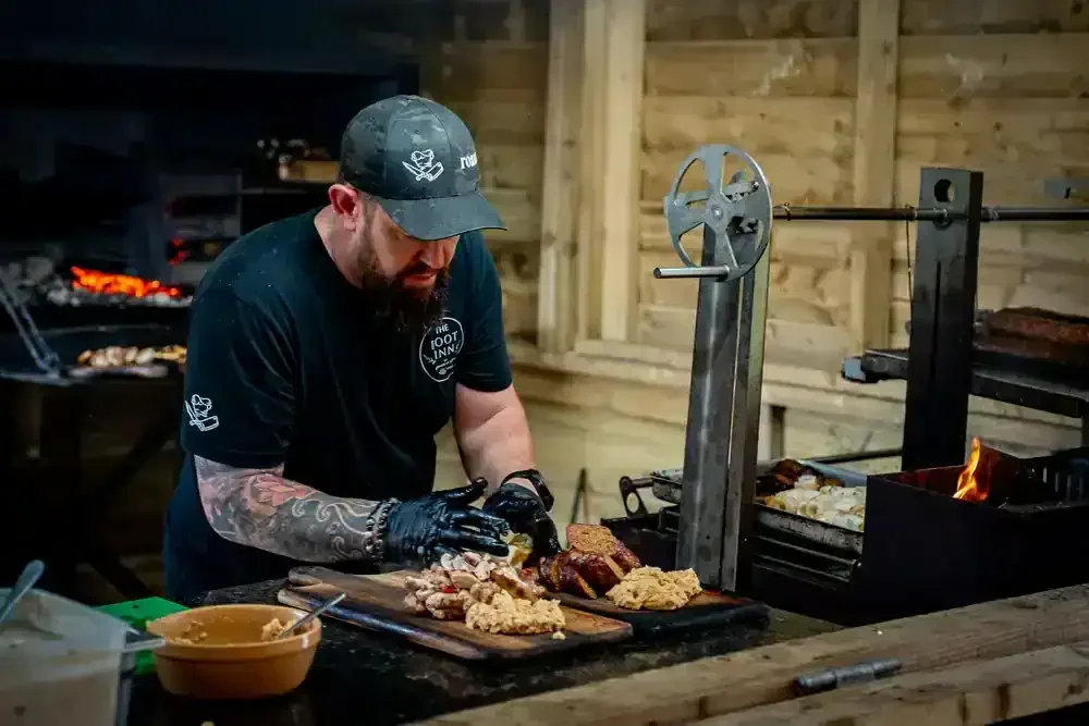 A man wearing a black cap and black gloves prepares fried chicken and beef brisket on a wooden countertop in a rustic kitchen or smoker area.