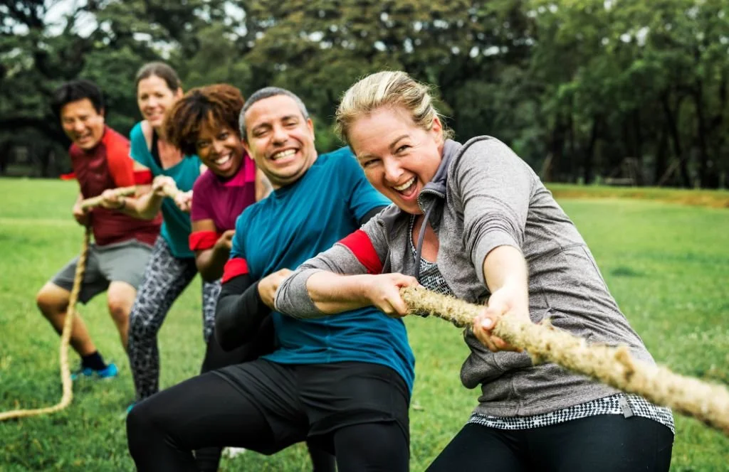 Group of five people playing tug-of-war outdoors on a grassy field, smiling and laughing, with trees in the background.