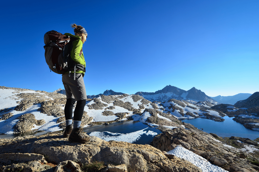 A hiker standing on a rocky mountain terrain with snow patches, looking at distant mountain peaks under a clear blue sky.