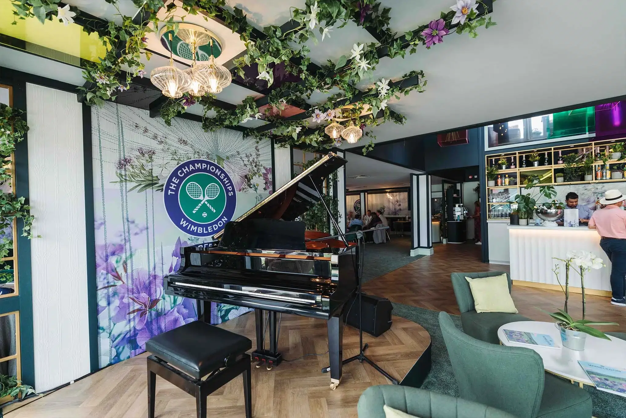 Interior of a restaurant or lounge with a grand piano, green chairs, a floral backdrop with Wimbledon logo, and a ceiling decorated with hanging greenery and flowers.