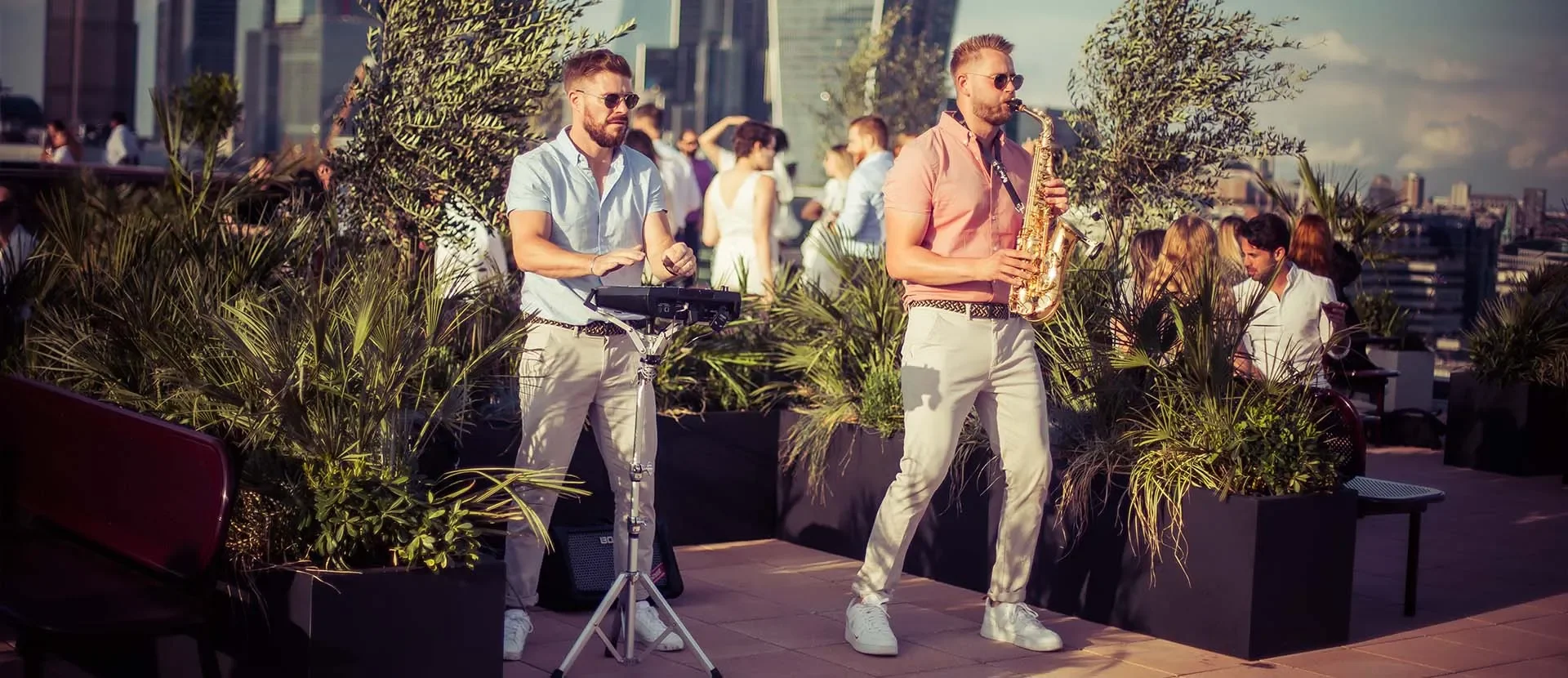 Two men performing live music on a rooftop terrace with city skyline in the background. One man is playing an electronic keyboard, and the other is playing a saxophone. They are dressed in light-colored clothes and wearing sunglasses. Several people are socializing in the background.