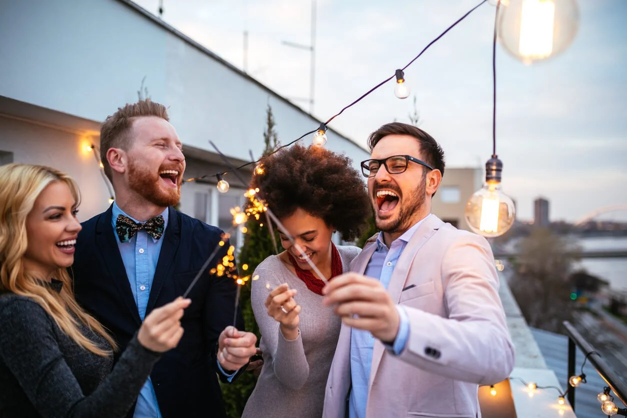 group of four friends celebrating on a rooftop at sunset, holding sparklers and smiling, with string lights hanging overhead.