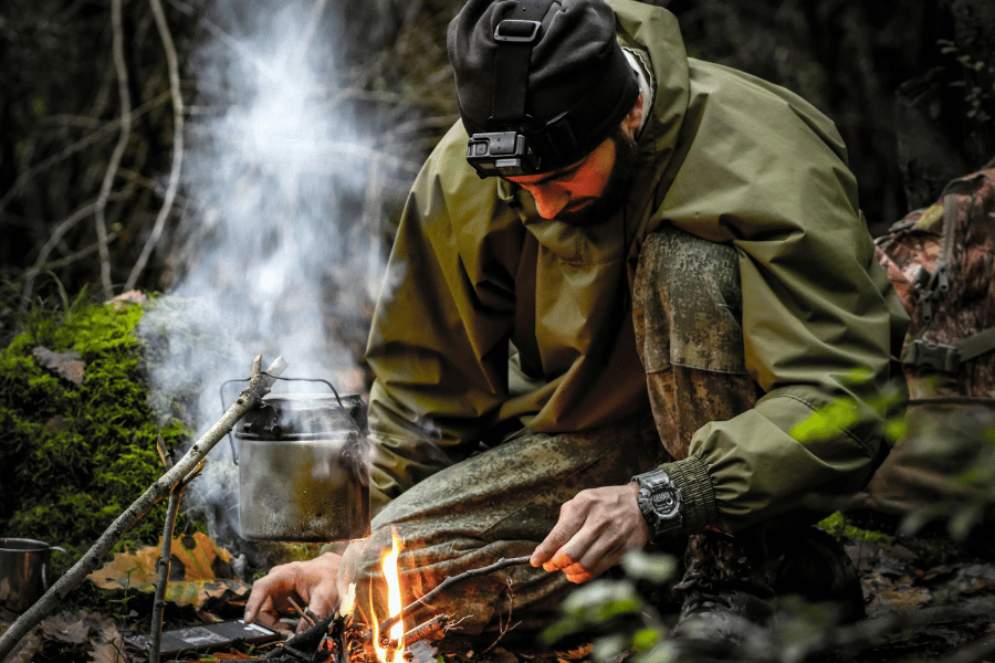 A man camping in the wilderness, tending to a small campfire with a pot hanging over it, surrounded by trees and greenery.