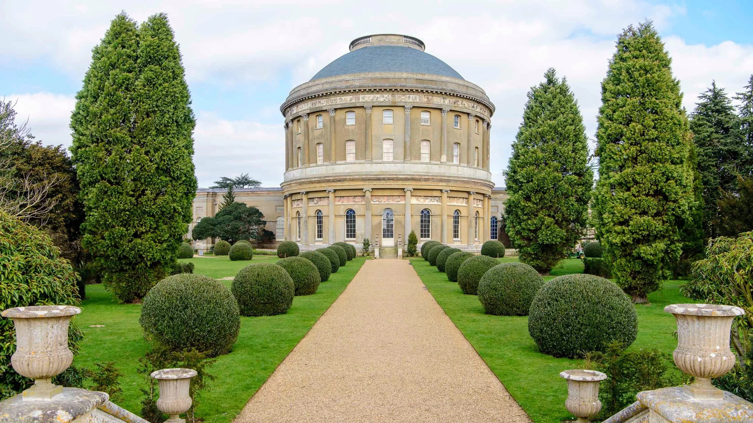 A large historic building with a rounded dome shape, surrounded by neatly trimmed bushes and green grass, with a gravel pathway leading to the entrance.